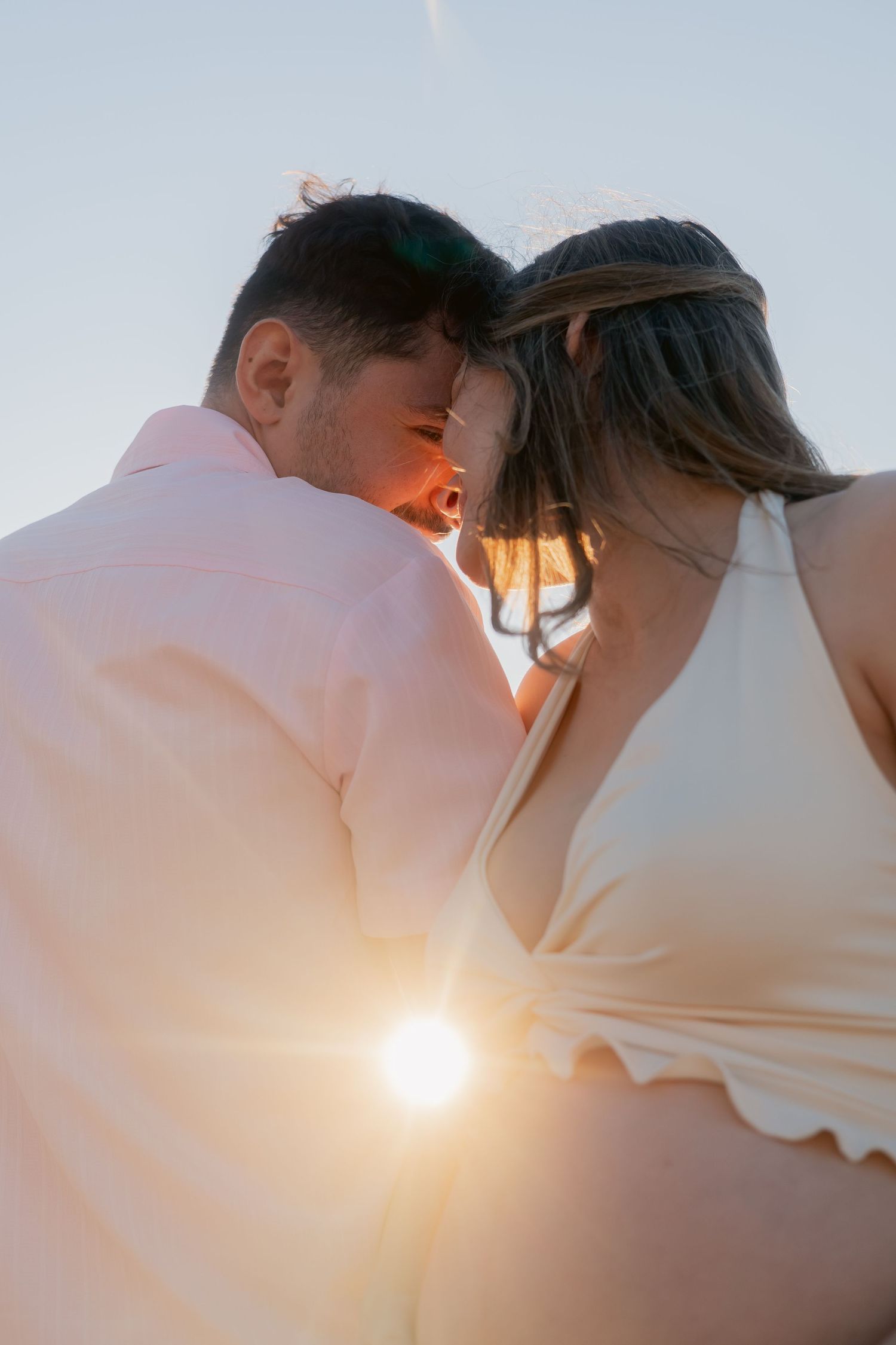 Close-up of couple touching foreheads at sunset, woman in white dress with floral headband, golden light.