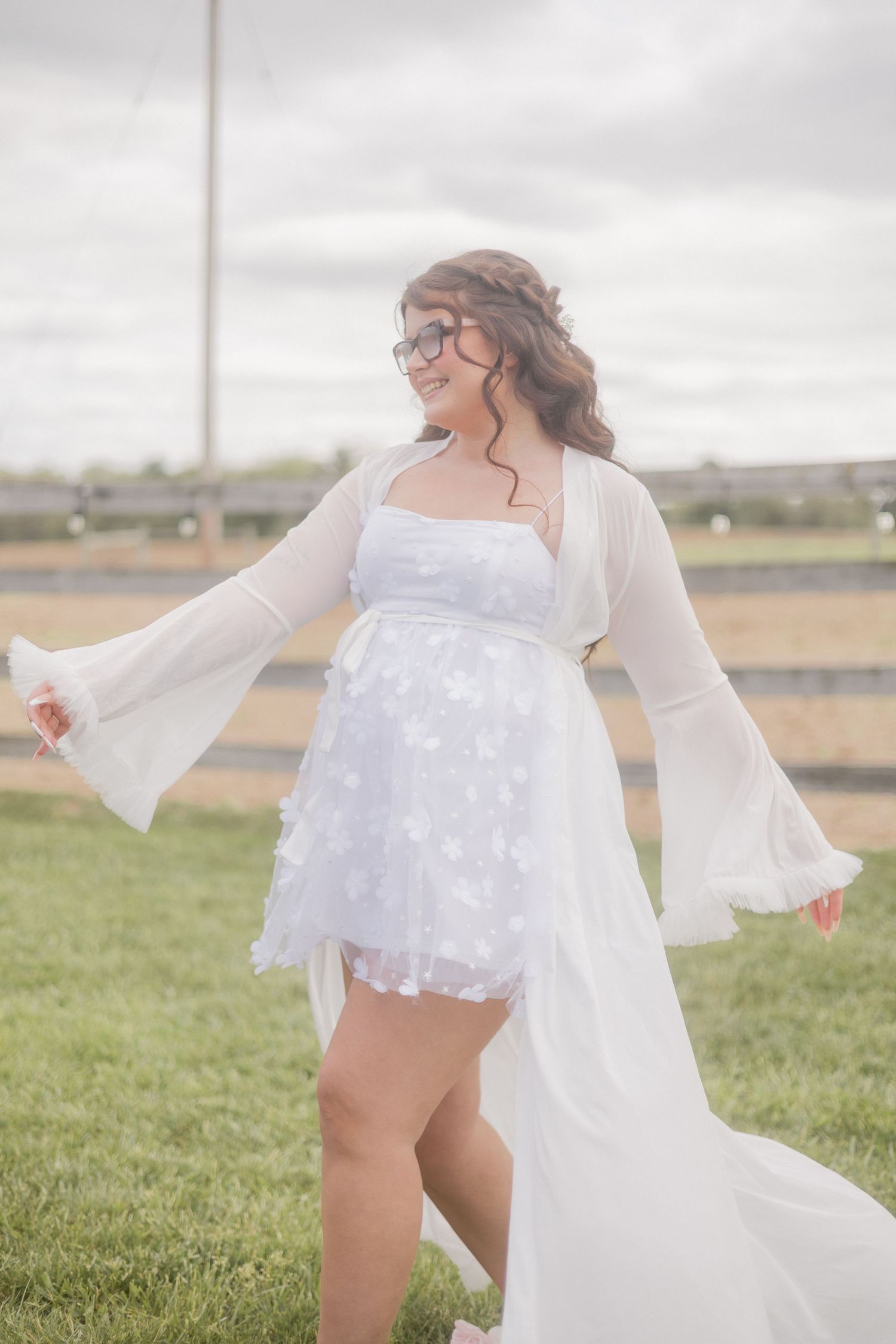 Smiling bride in white floral mini dress with sheer bell sleeves spinning joyfully on green lawn.