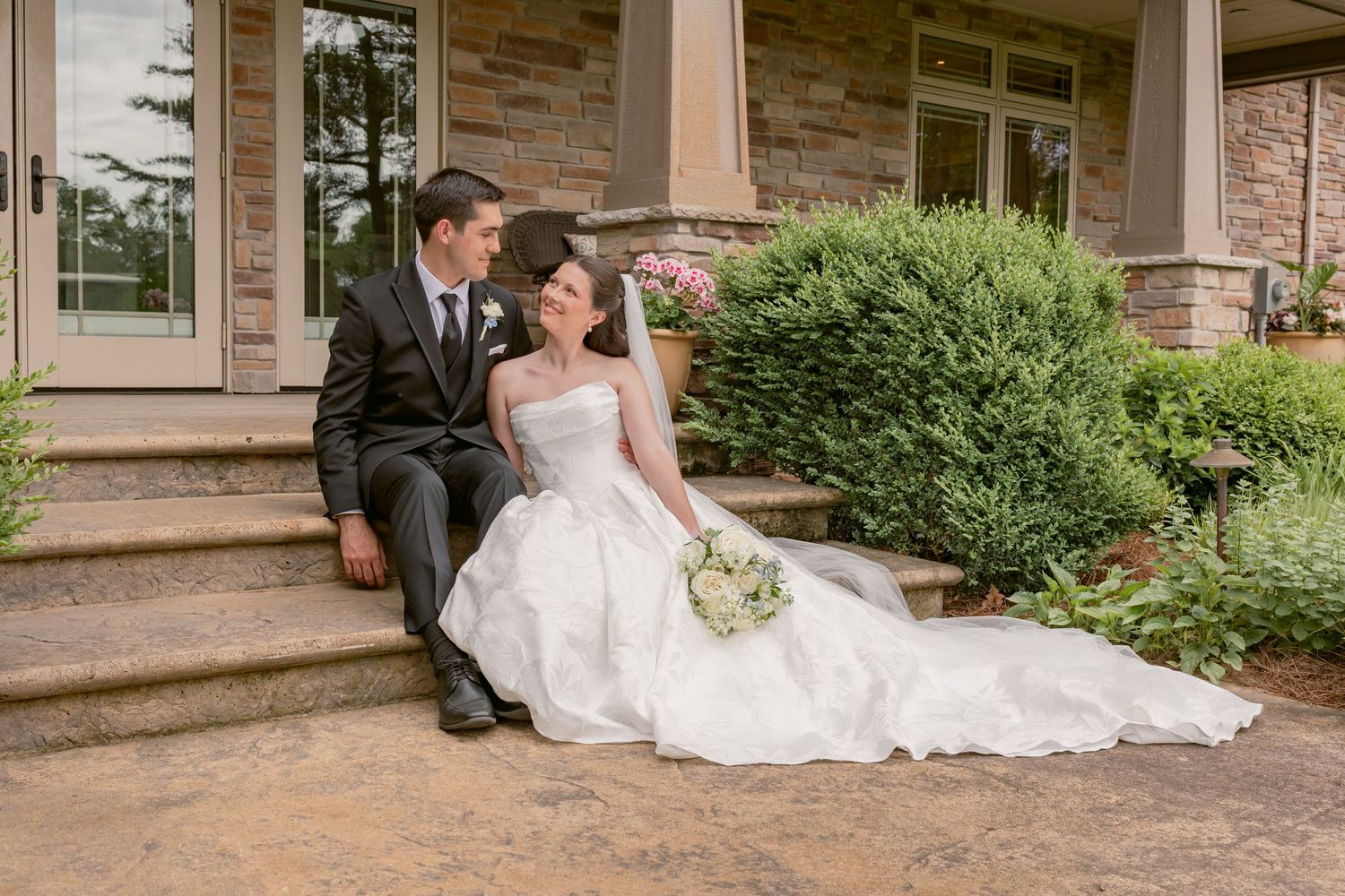 Bride and groom sitting on stone steps outside a elegant home, sharing a romantic moment on their wedding day.