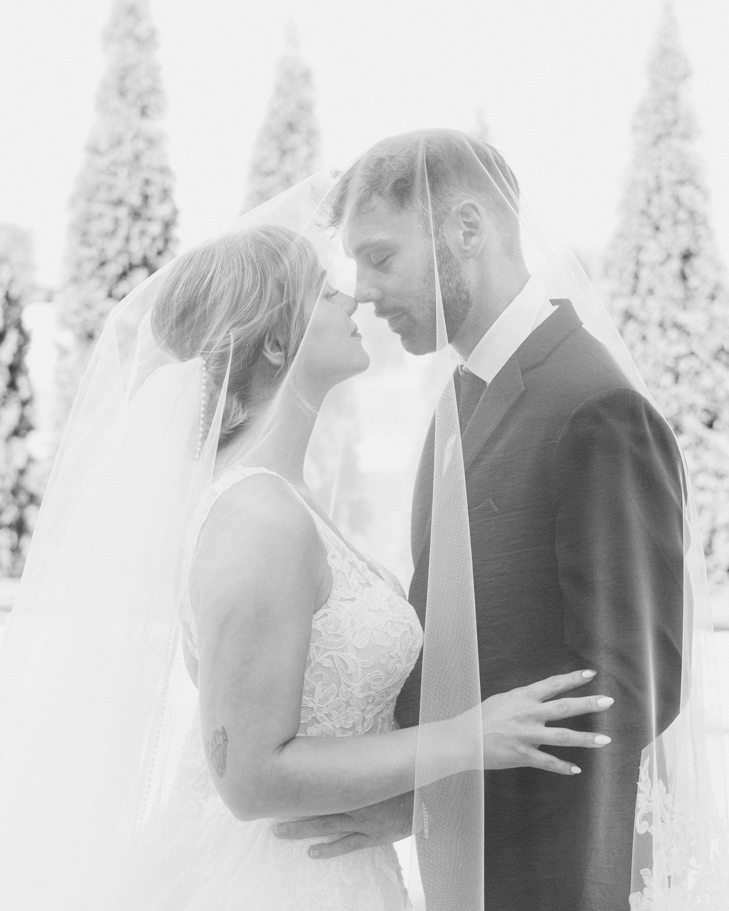 Black and white romantic wedding portrait of bride and groom under flowing veil outdoors.