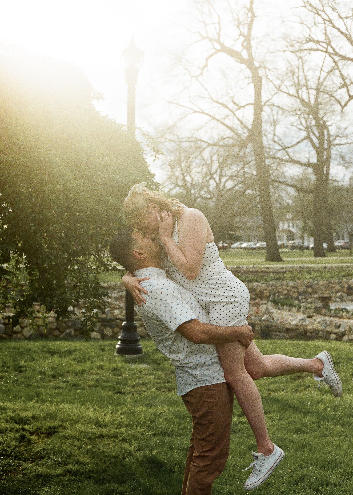 Man lifting woman for a kiss in sunlit park, woman wearing white polka dot dress and sneakers.