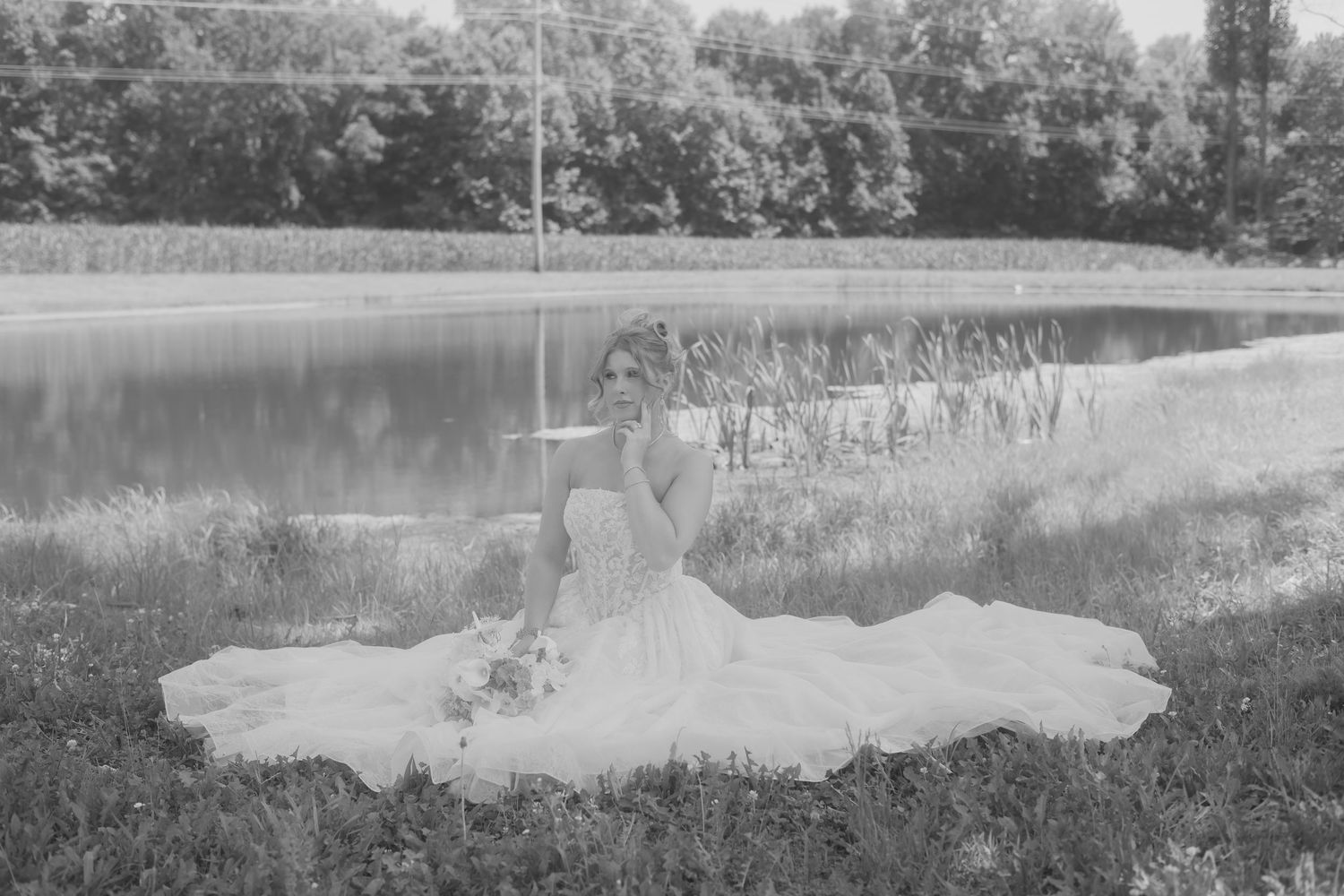 Black and white photo of a bride in a flowing gown sitting gracefully on grass beside a calm pond.