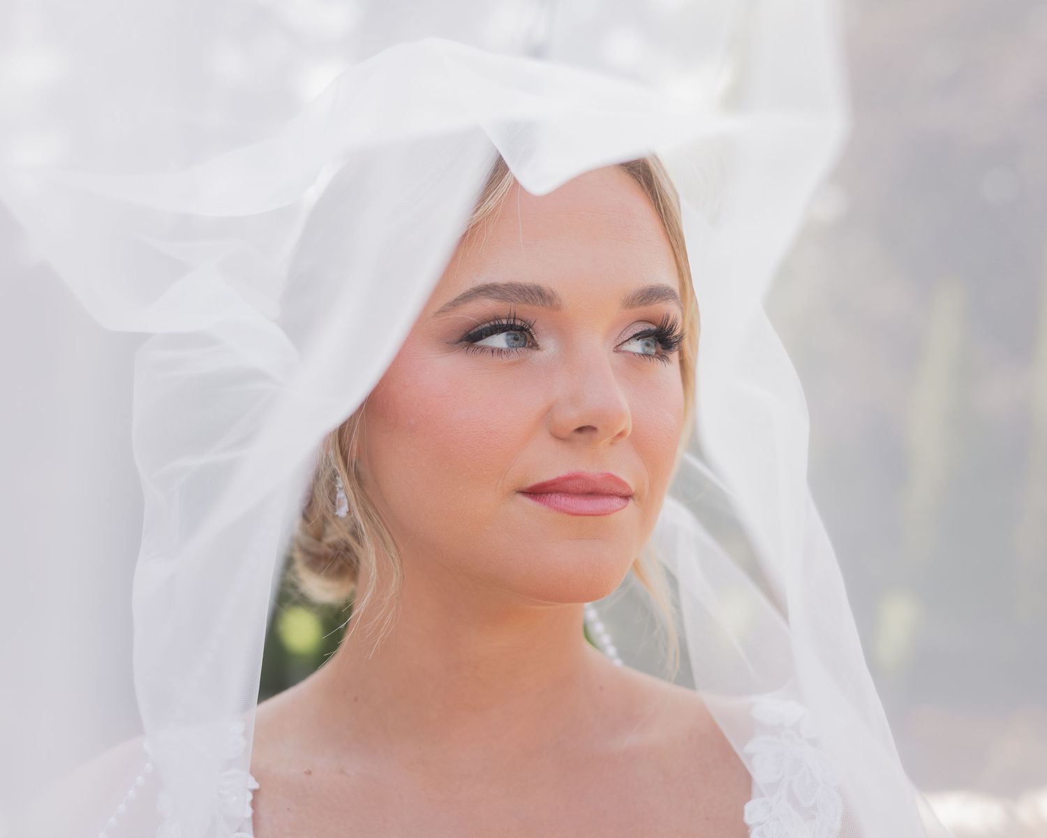 Close-up portrait of beautiful bride wearing white veil with bow, natural glam makeup outdoors.