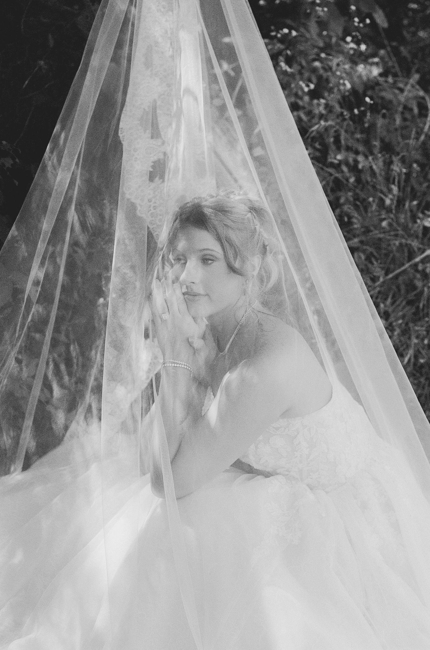 Black and white photo of a bride in a lace wedding gown beneath a flowing veil outdoors.