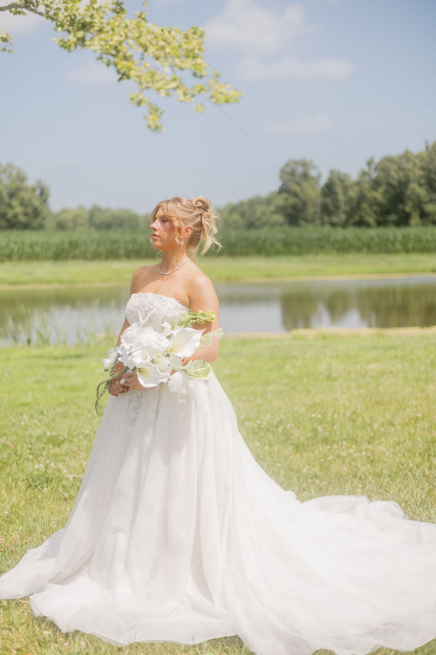Elegant bride holding white orchid bouquet stands in flowing strapless wedding gown beside a peaceful pond.