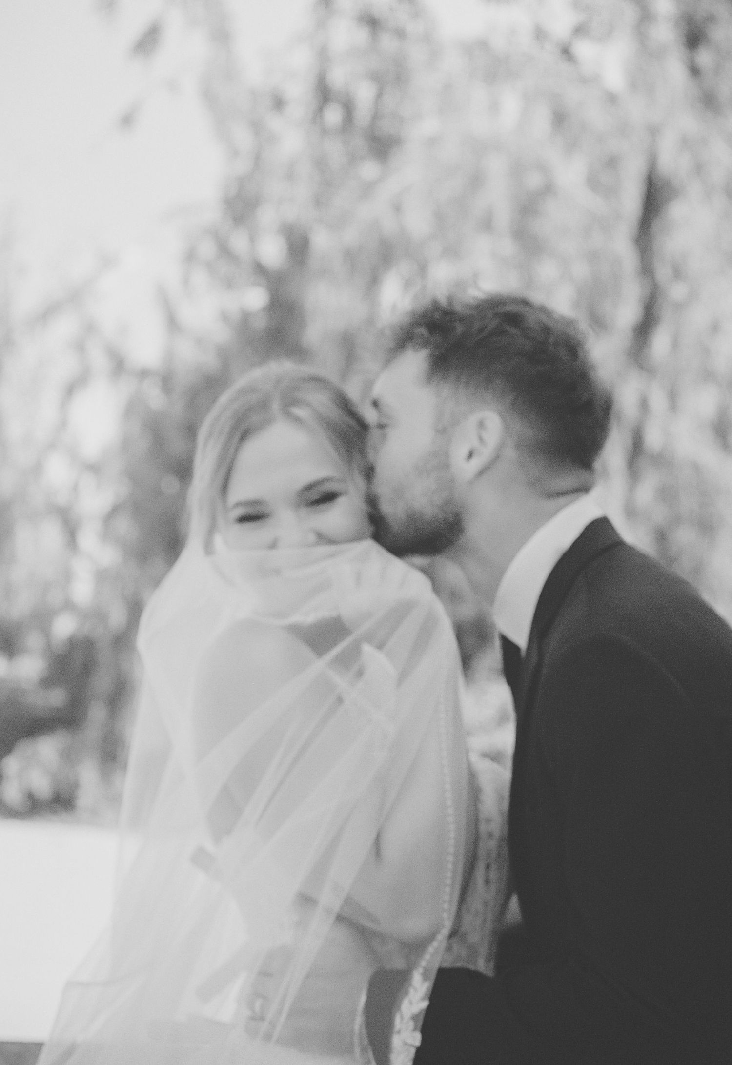Black and white photo of groom kissing bride on cheek as she smiles beneath flowing wedding veil.