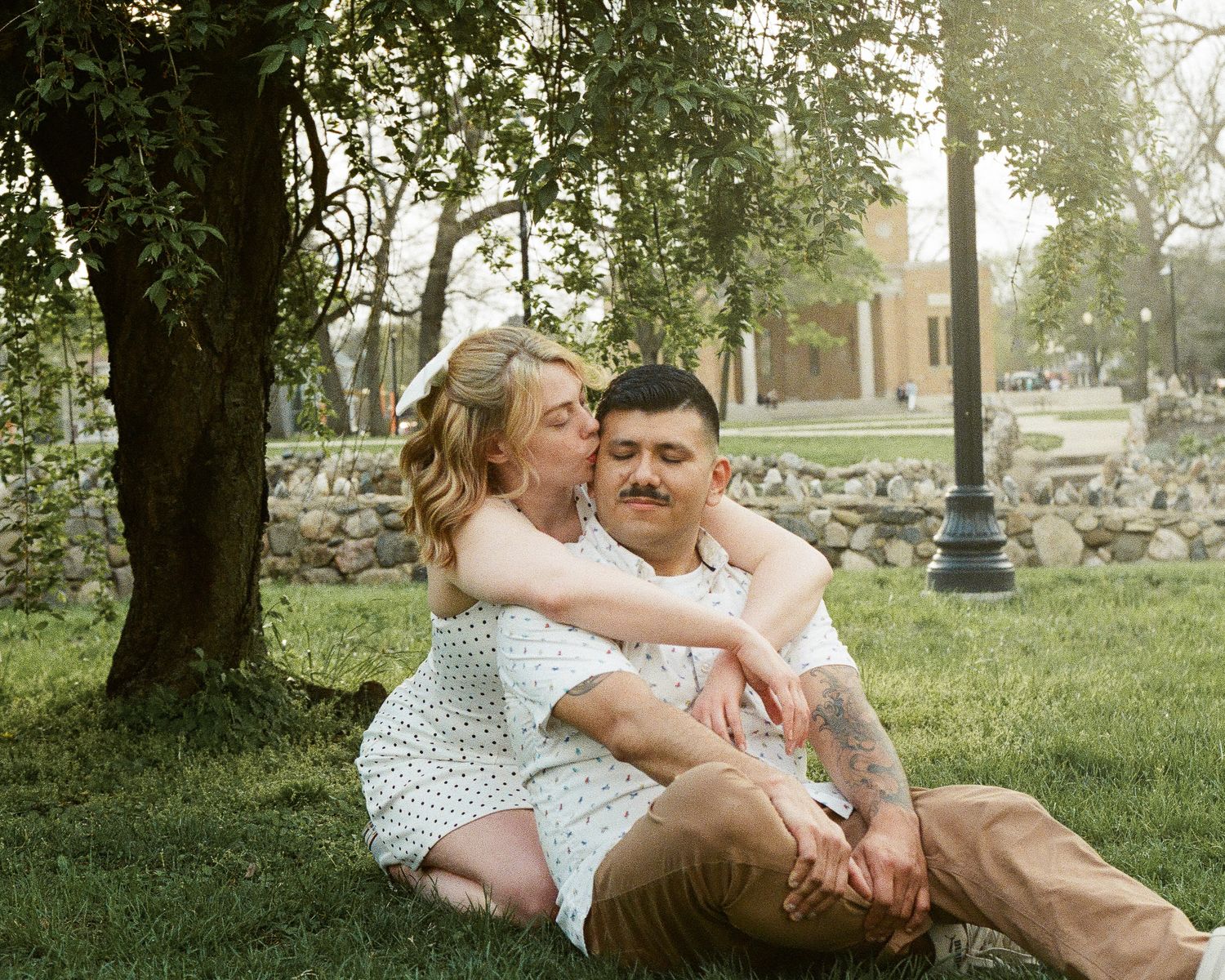 Couple sitting together on green grass under a tree, woman in white dress embracing man with tattoos outdoors.