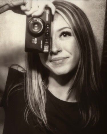 Young woman smiling while holding a vintage camera up to her eye in black and white.