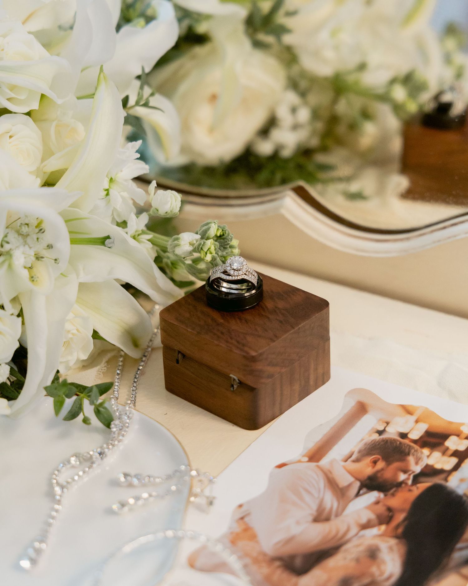 Wedding rings displayed in walnut ring box beside white roses and romantic couple photo.