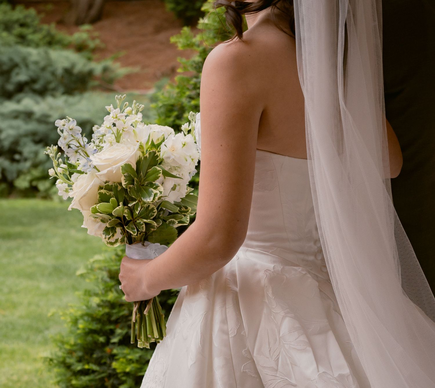 Back view of a bride in a strapless white gown holding a white floral bouquet in a garden setting.