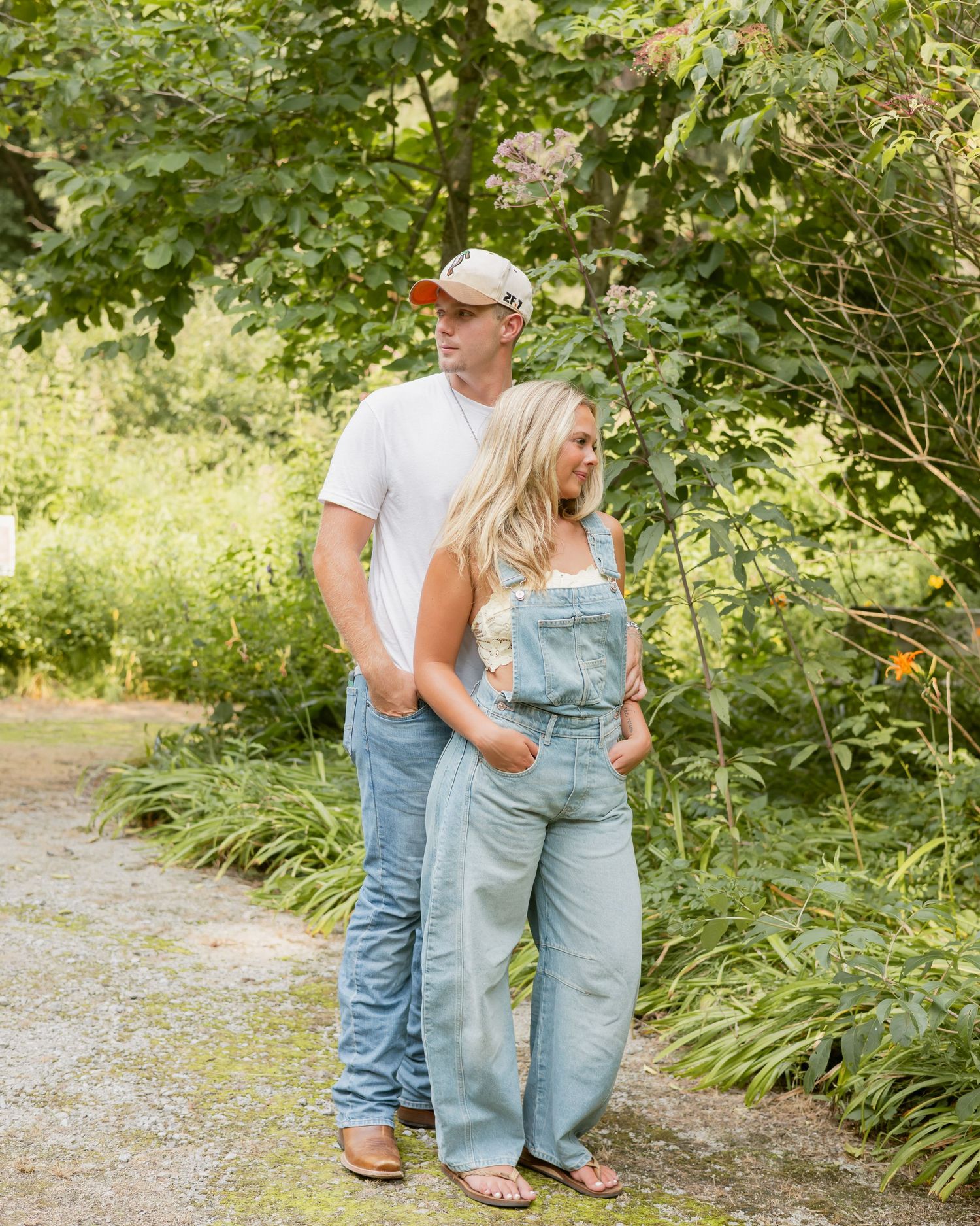 Tall man and blonde woman in denim overalls pose together on garden path among greenery.
