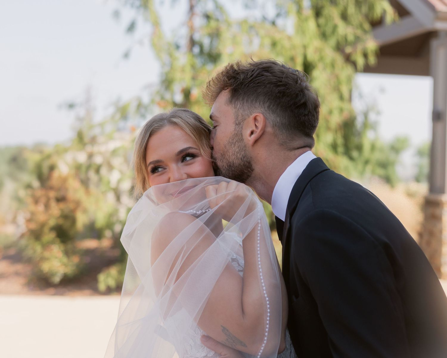 Groom tenderly kisses blonde bride on the cheek while her veil covers her smiling face.