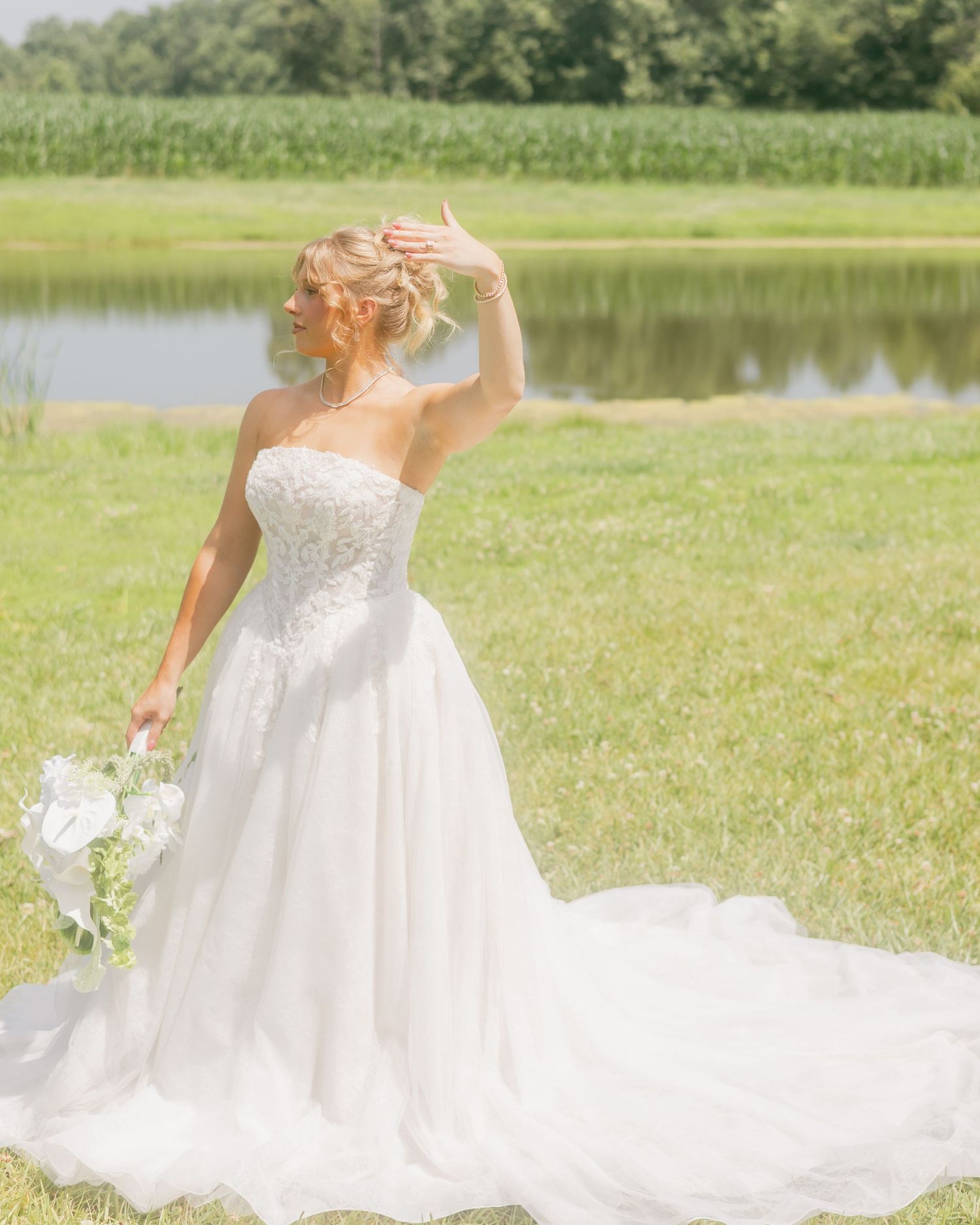 Bride in strapless white ballgown with lace bodice poses gracefully by a serene pond on a sunny day.