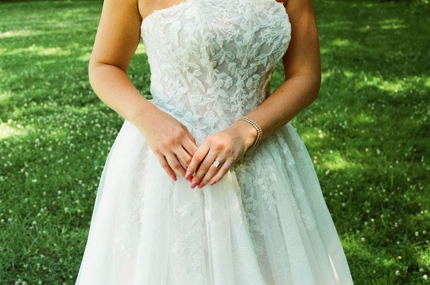 Close-up of bride in strapless lace wedding gown with red manicured nails and gold rings on green grass background.