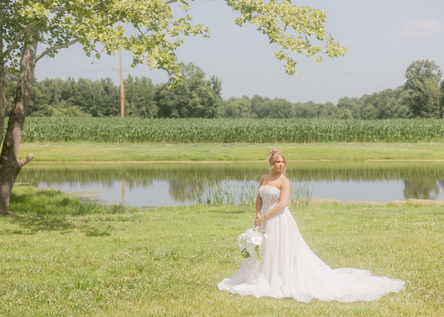 Blonde bride in a strapless white ballgown with long train poses near a tranquil pond surrounded by greenery.