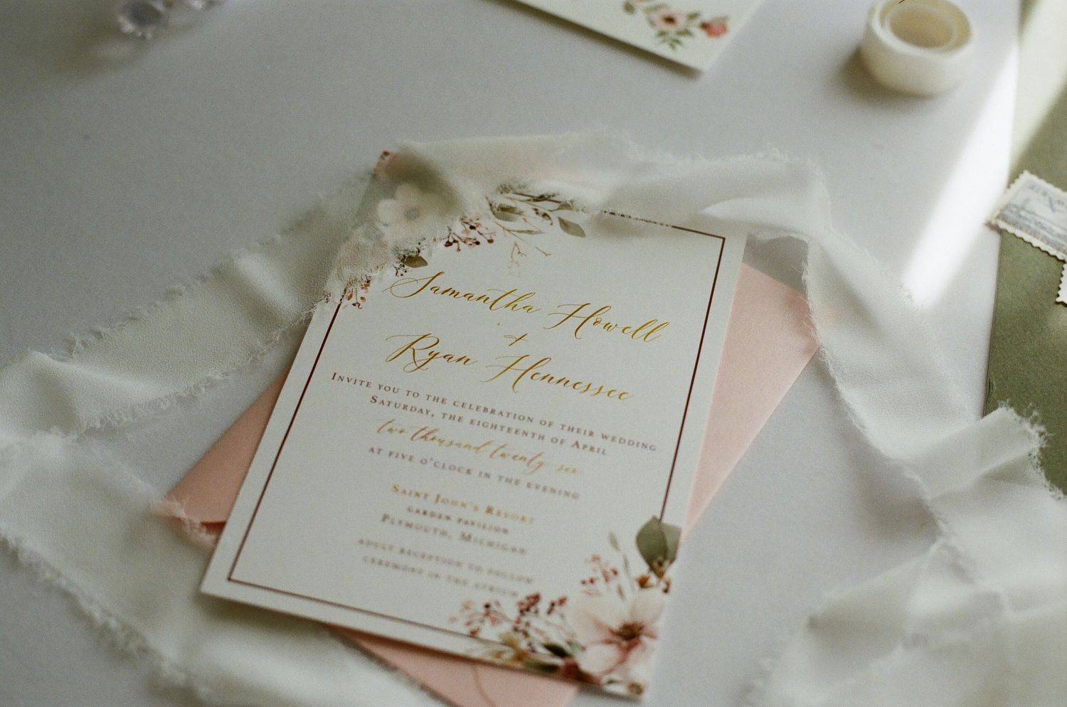 Elegant gold foil wedding invitation with floral watercolor accents on blush pink envelope with white ribbon details.