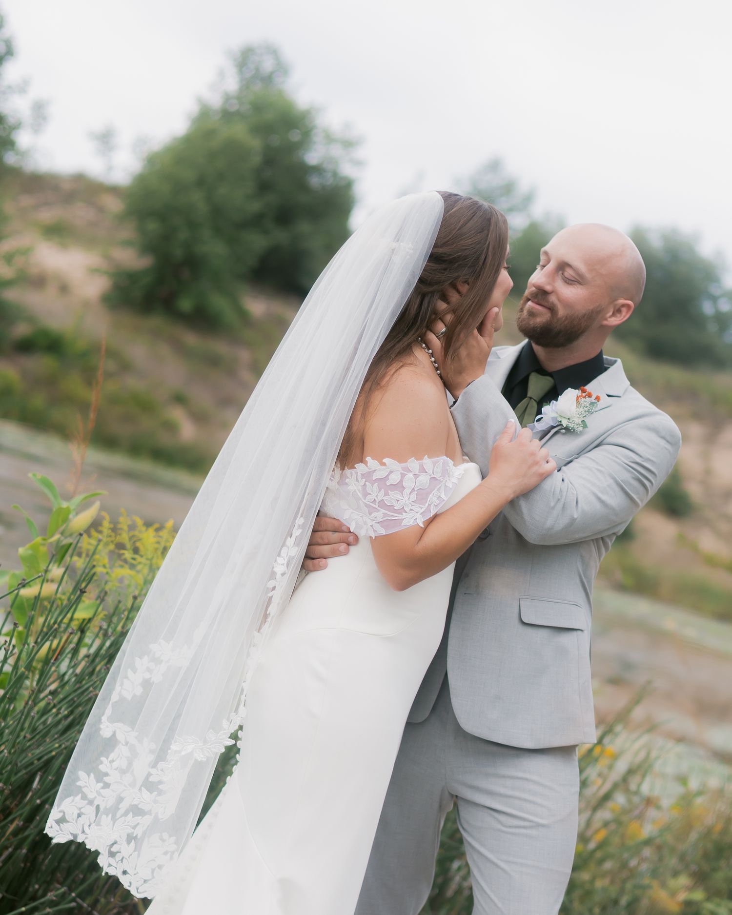 Bald groom in grey suit gazes lovingly at bride in lace off-shoulder gown with long veil outdoors.