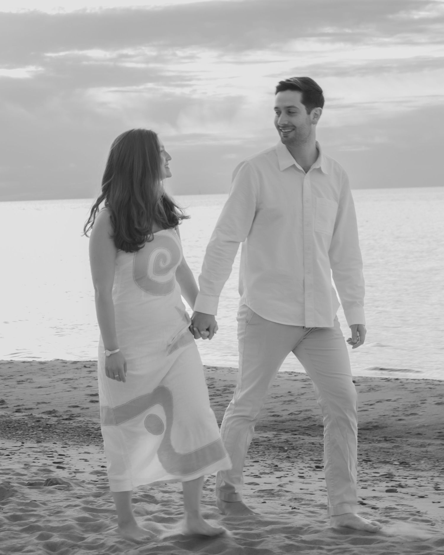 Black and white photo of a couple holding hands and walking barefoot on a sandy beach.