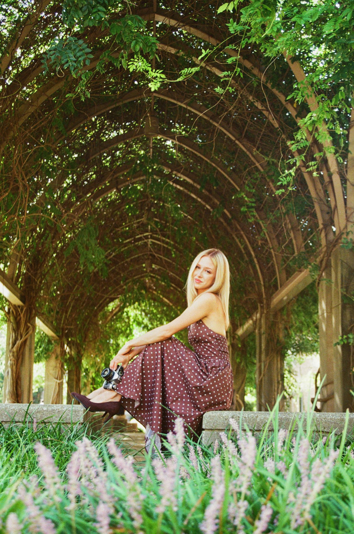 Blonde woman in a polka dot maxi dress sitting under a lush green arched garden tunnel.