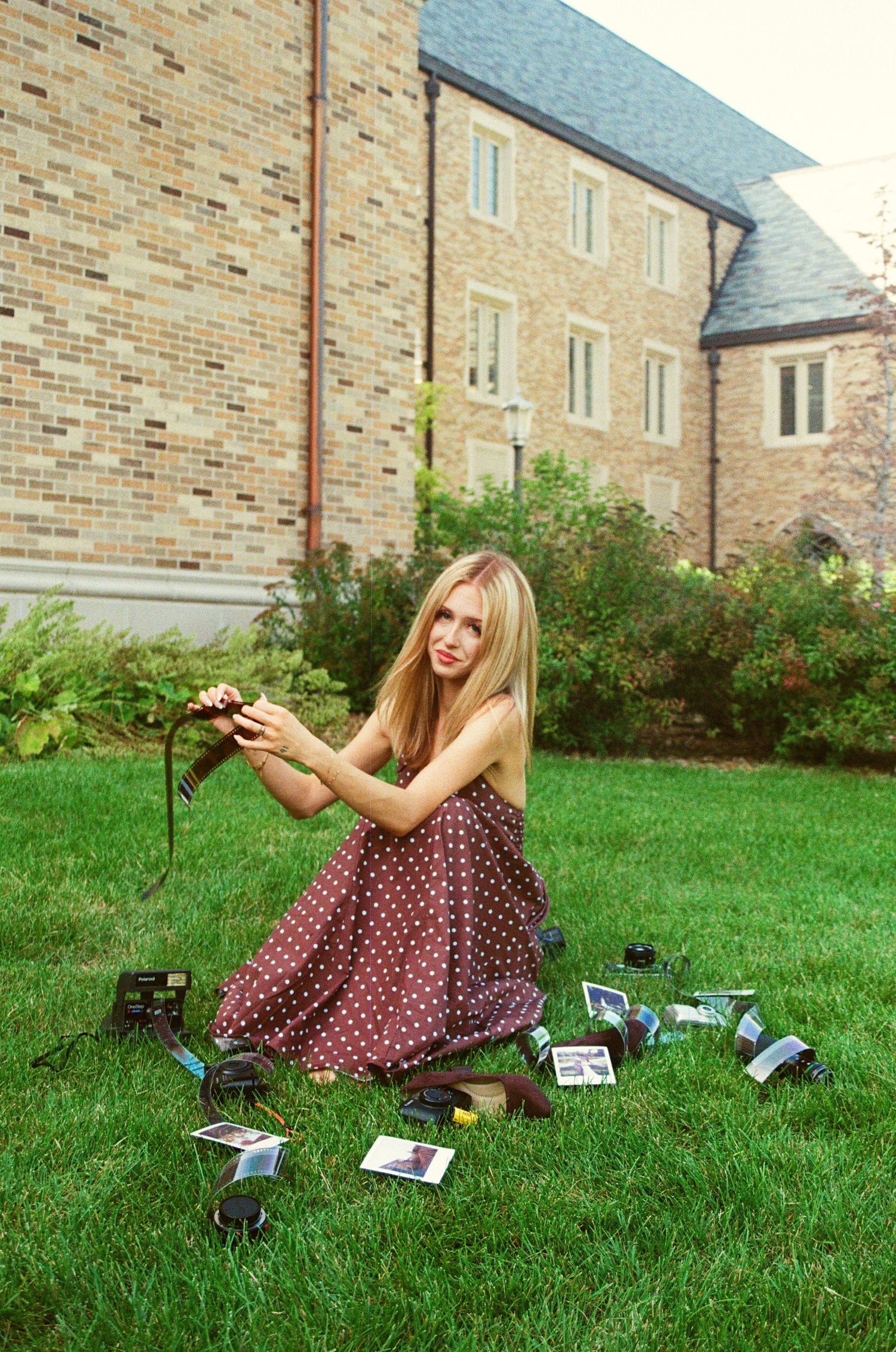 Blonde woman in polka dot dress kneeling on grass surrounded by polaroid photos and cameras outside brick building.