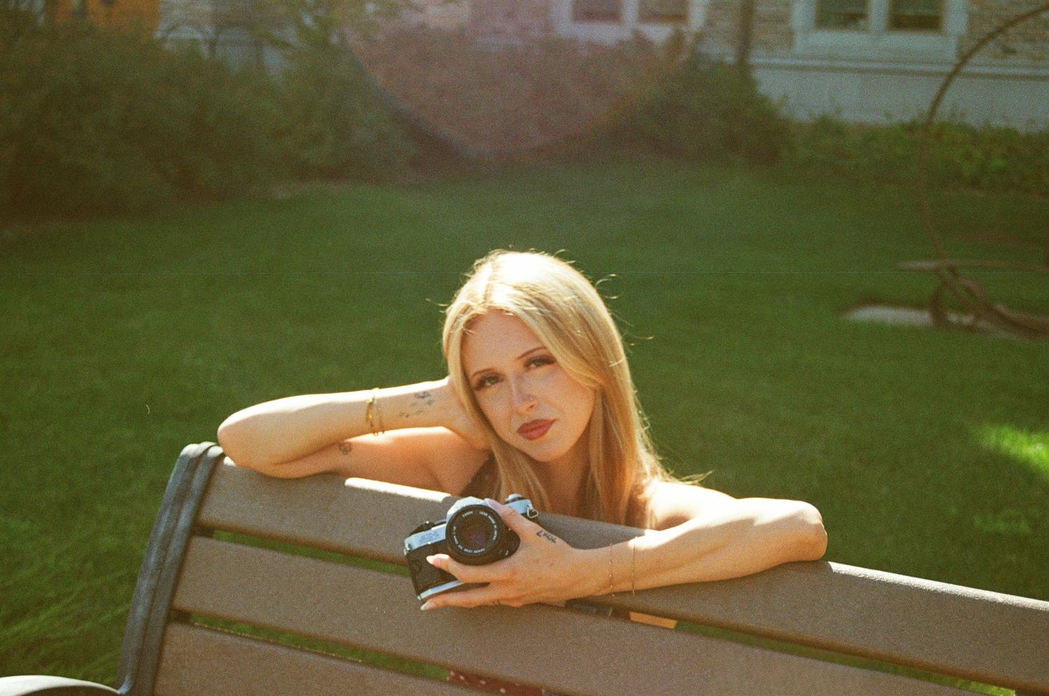 Blonde woman leaning on a wooden park bench in golden sunlight holding a film camera.