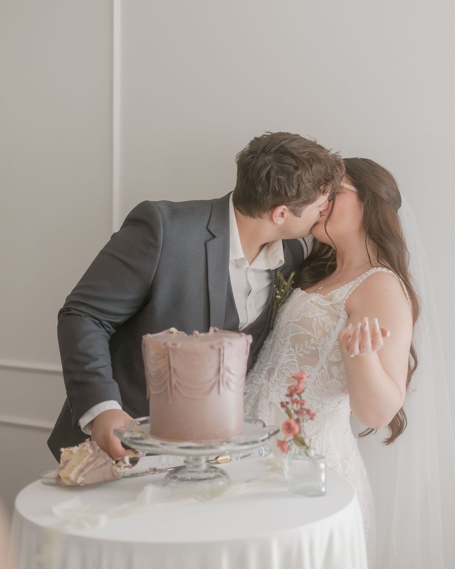 Newlyweds sharing a kiss while cutting elegant pink wedding cake at white reception table.