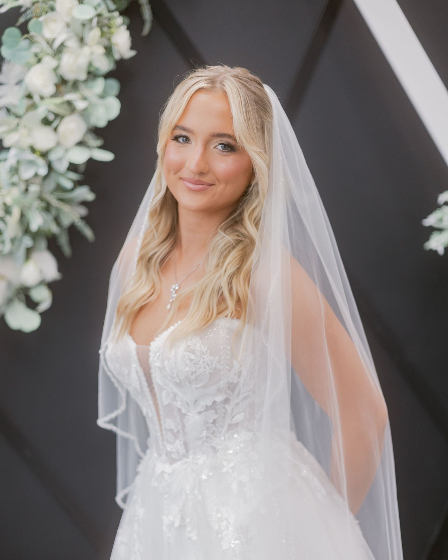 Blonde bride wearing a lace strapless gown and flowing veil smiles softly beneath a floral wedding arch.