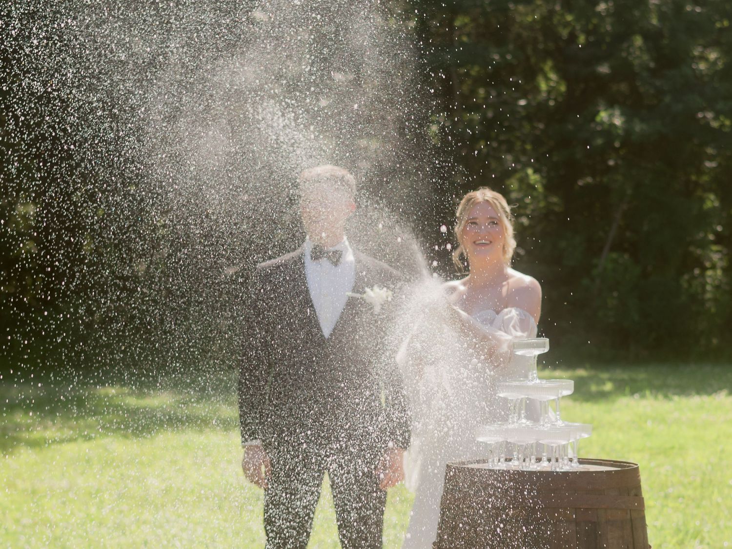 Joyful bride and groom celebrate with champagne tower fountain spraying outdoors on sunny wedding day.