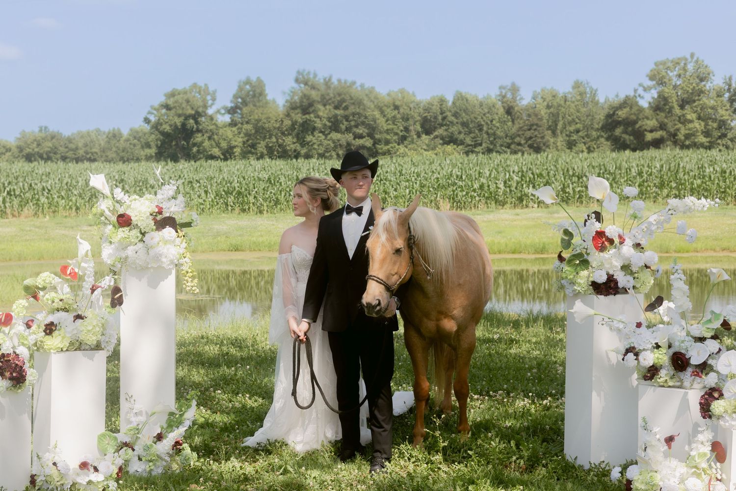 Bride and groom posing with a horse beside floral arrangements near a pond.