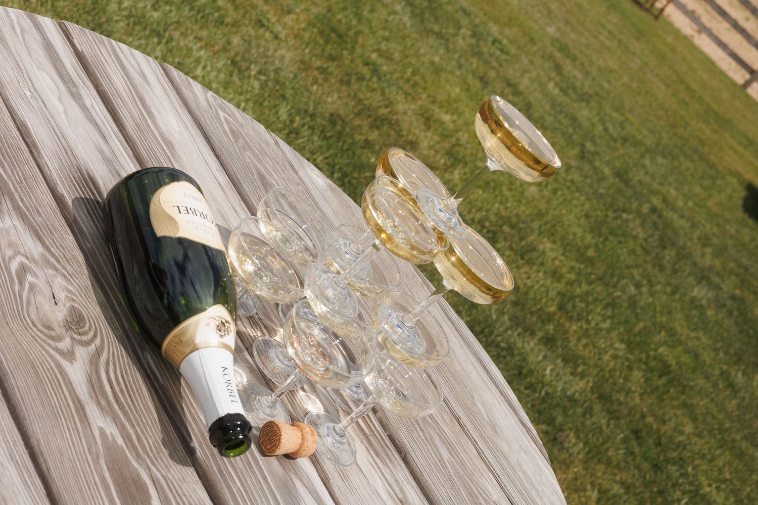 Champagne bottle and gold-rimmed glasses arranged on weathered wooden table on green lawn.