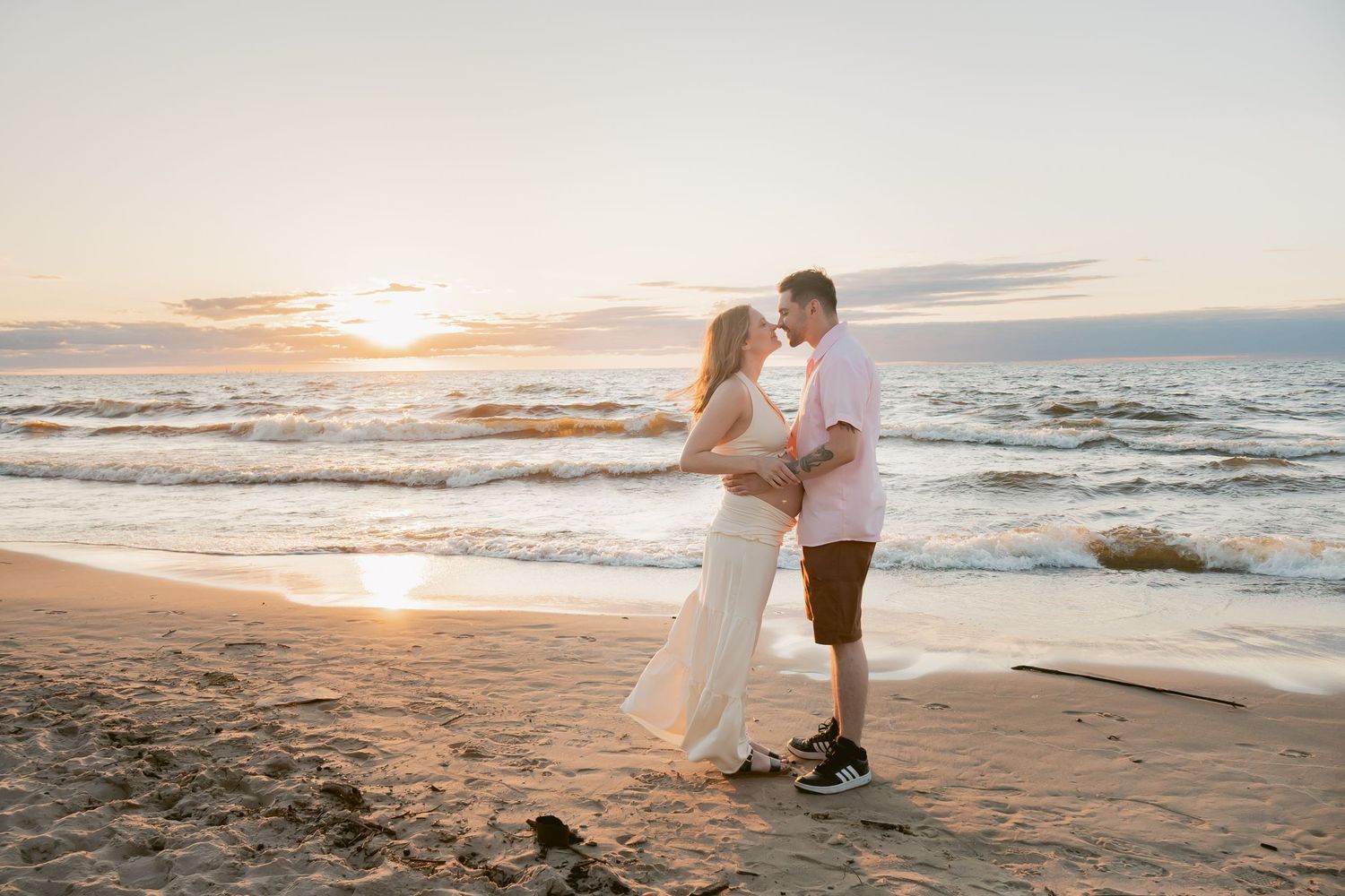 Pregnant couple embracing on sandy beach at sunset, woman in white dress, waves crashing behind them.