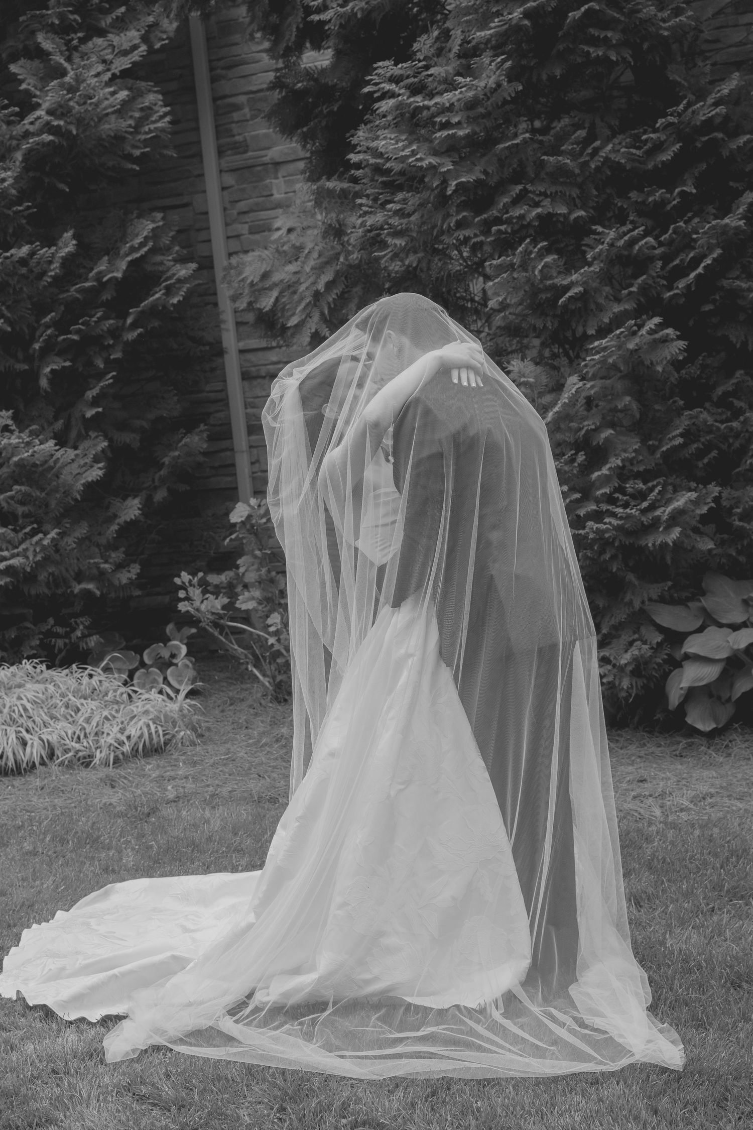 Artistic black and white photo of a bride covered by her flowing veil in a lush garden setting.