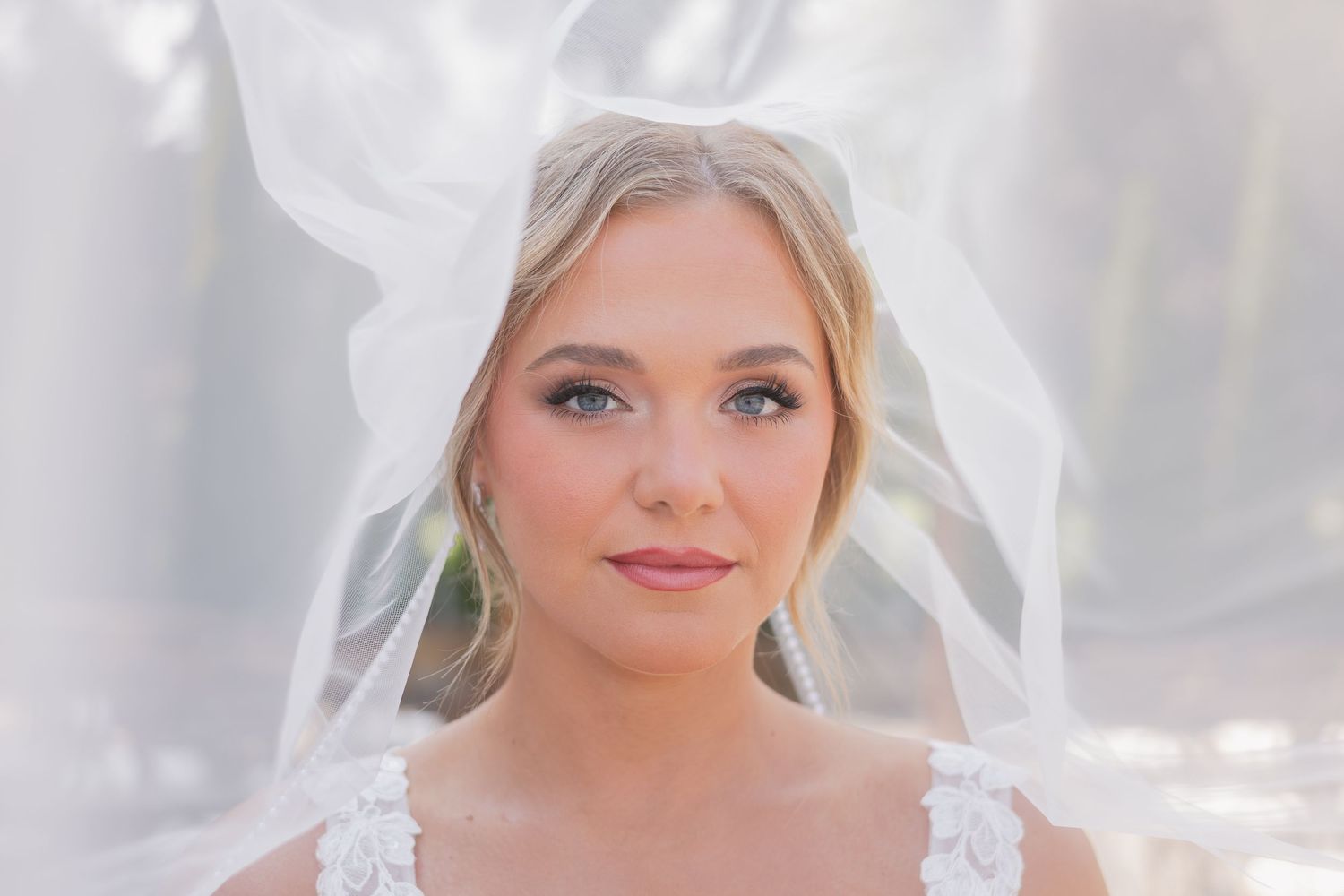 Beautiful blonde bride with blue eyes and rosy makeup gazing forward, draped in a flowing white wedding veil.