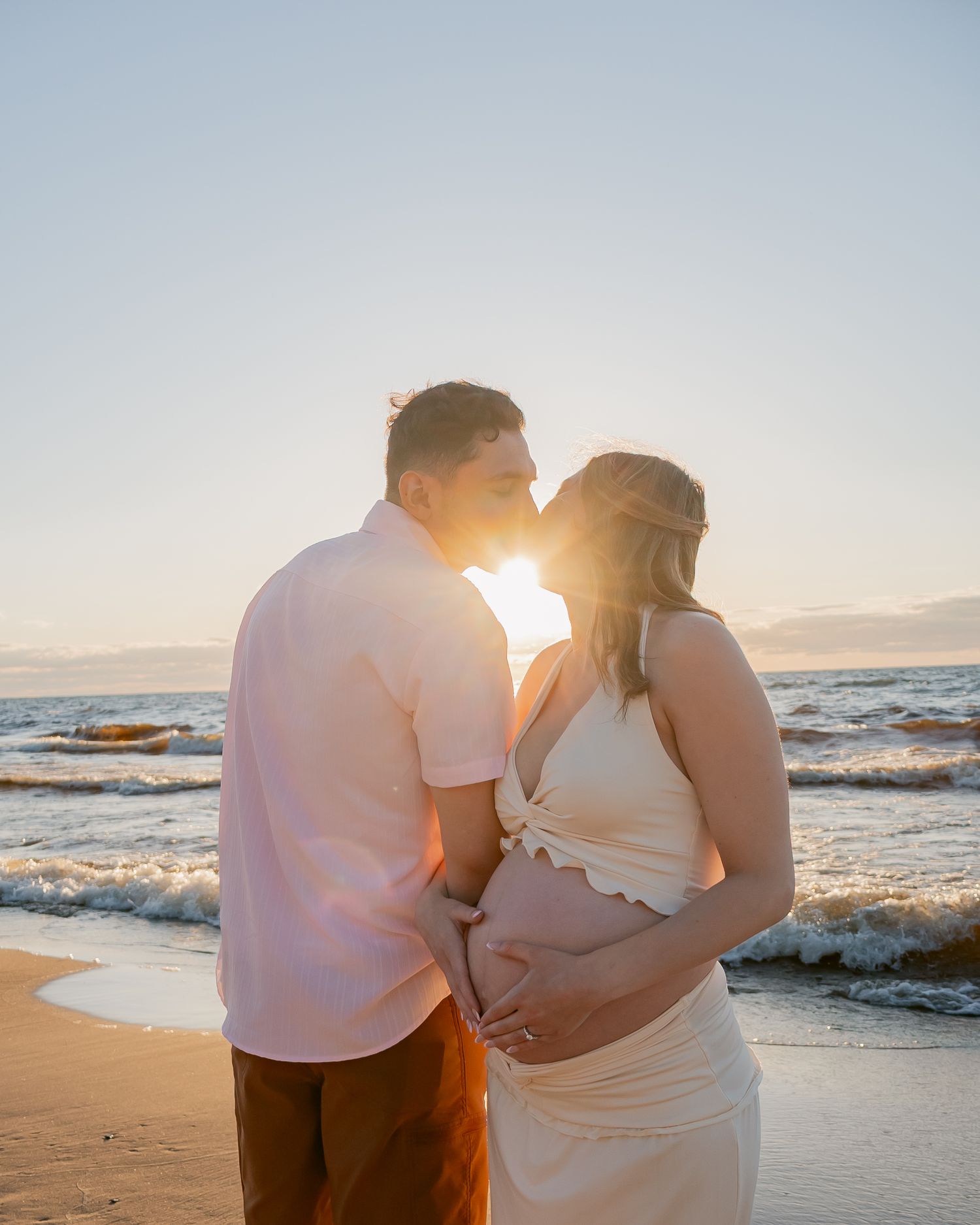 Couple kissing on beach at sunset while cradling pregnant belly with ocean waves behind them.