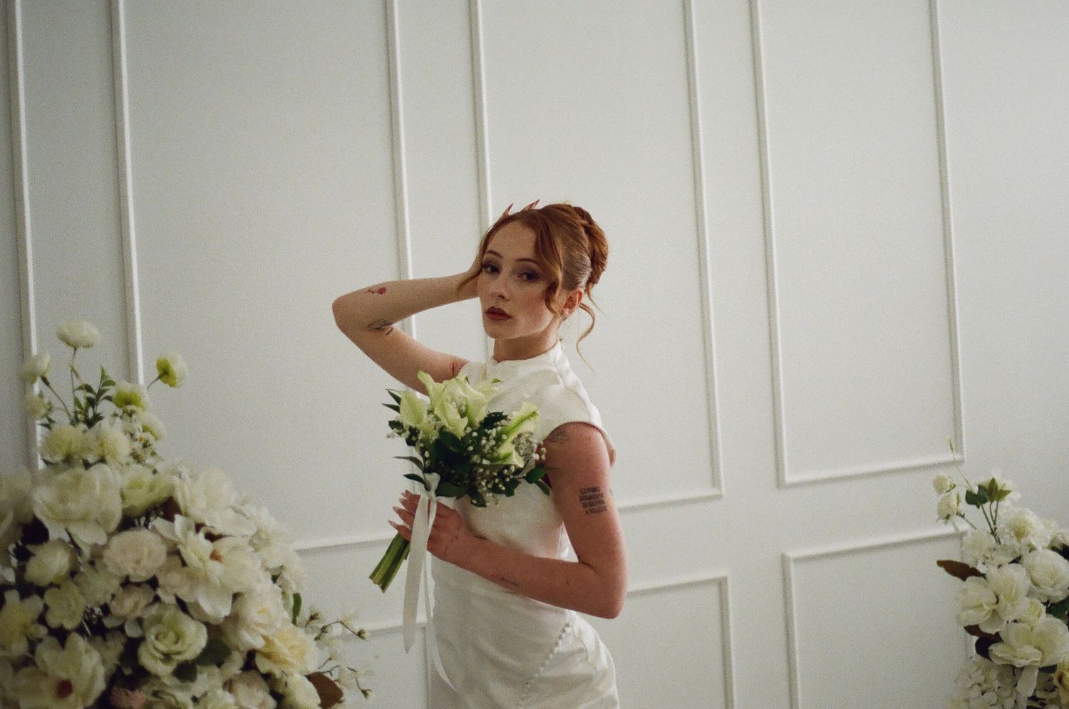 Bride in white gown holding bouquet of white flowers poses elegantly against white paneled wall.