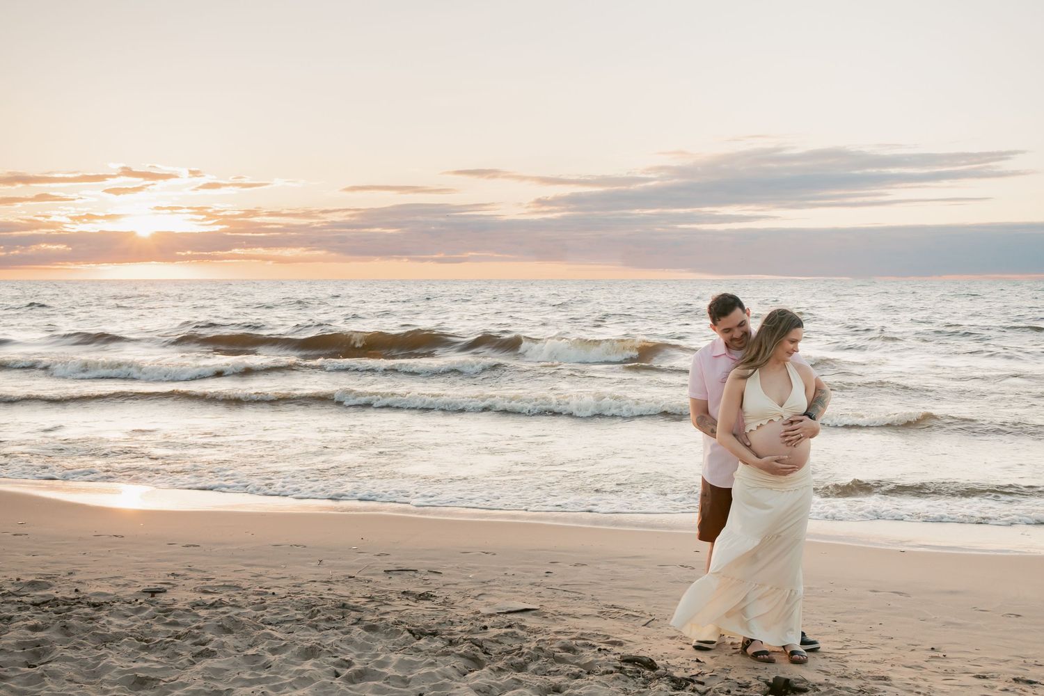 Couple walking on beach at sunset, man embracing pregnant woman in white dress, colorful sky behind them.