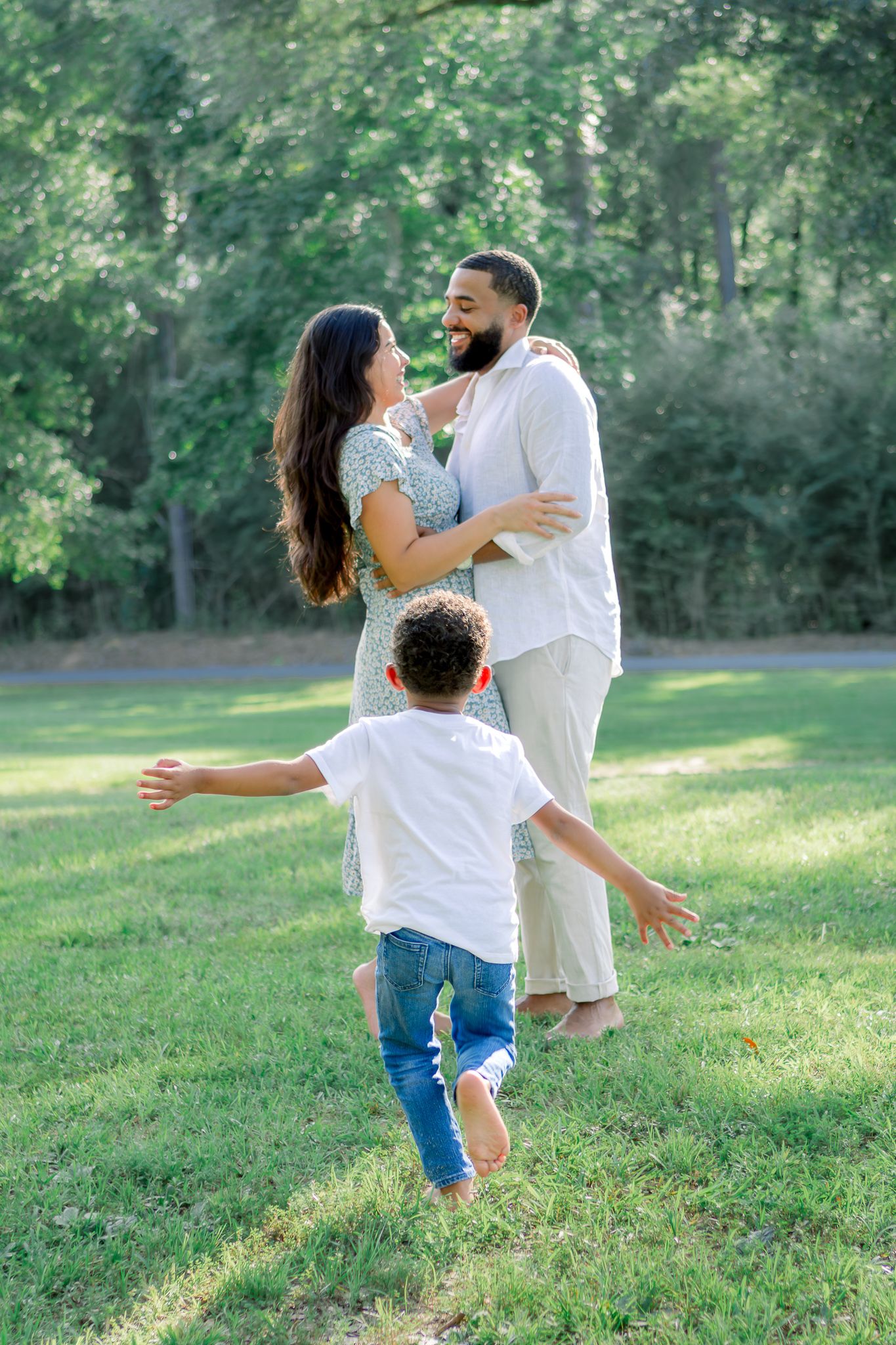 A joyful family in a sunny park, child running toward embracing parents on green grass.
