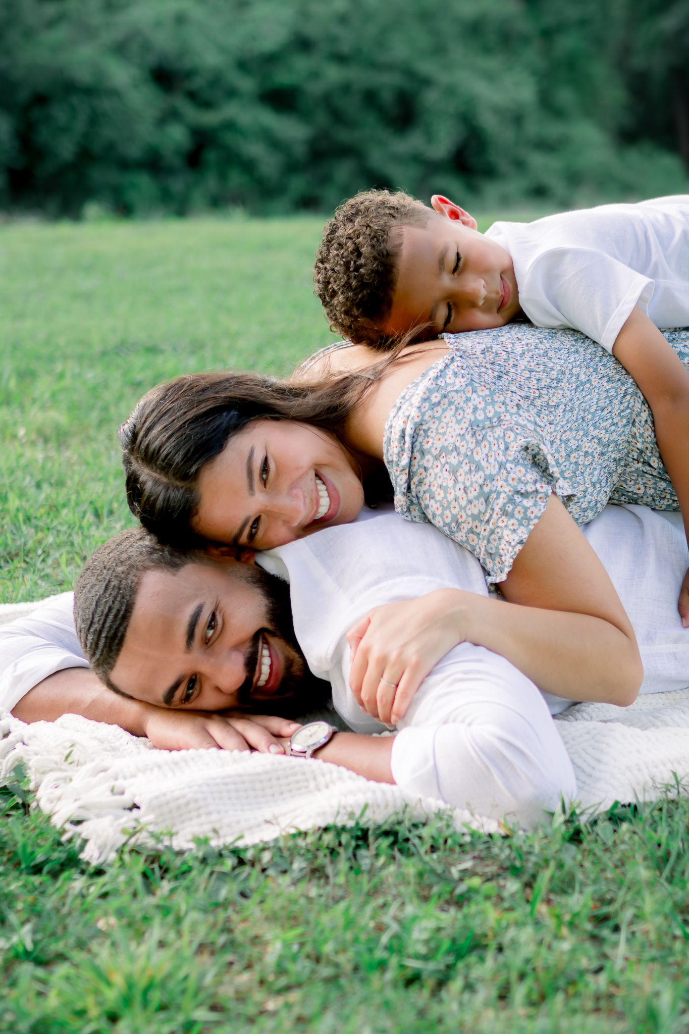 Joyful family of three lying on a blanket in the grass, smiling and stacking on top of each other.