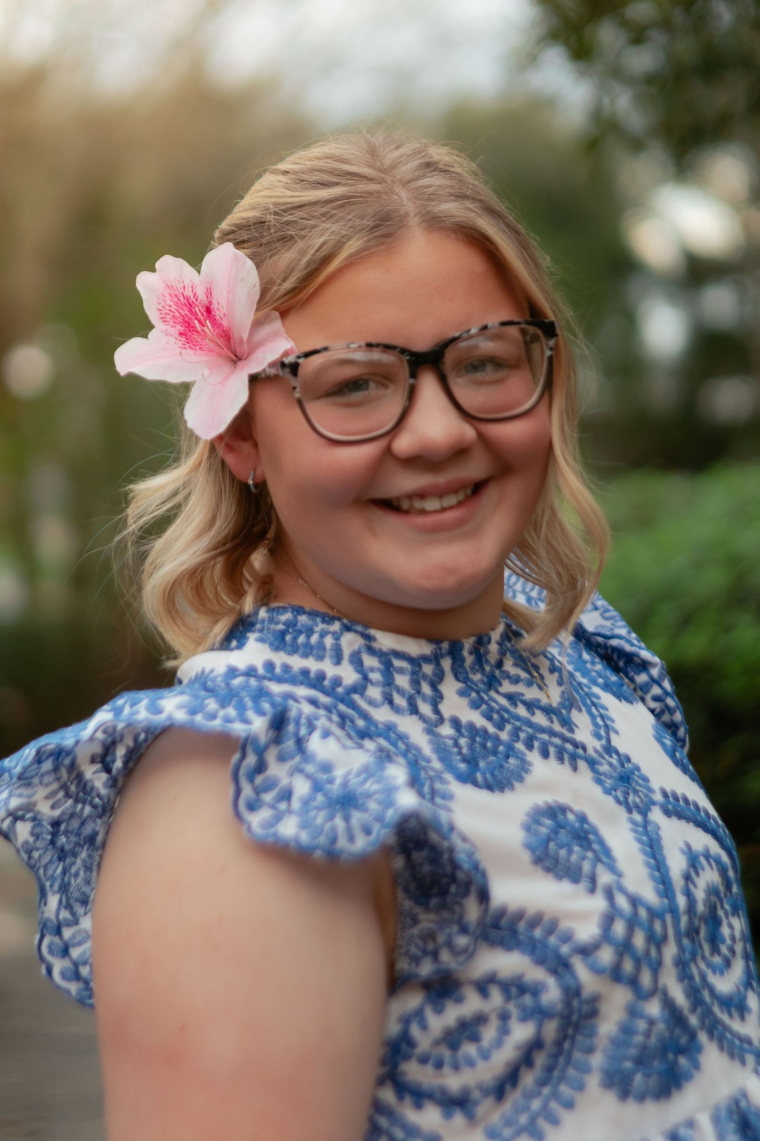 A person wearing glasses and a blue and white patterned dress smiles with a pink flower in their blonde hair.