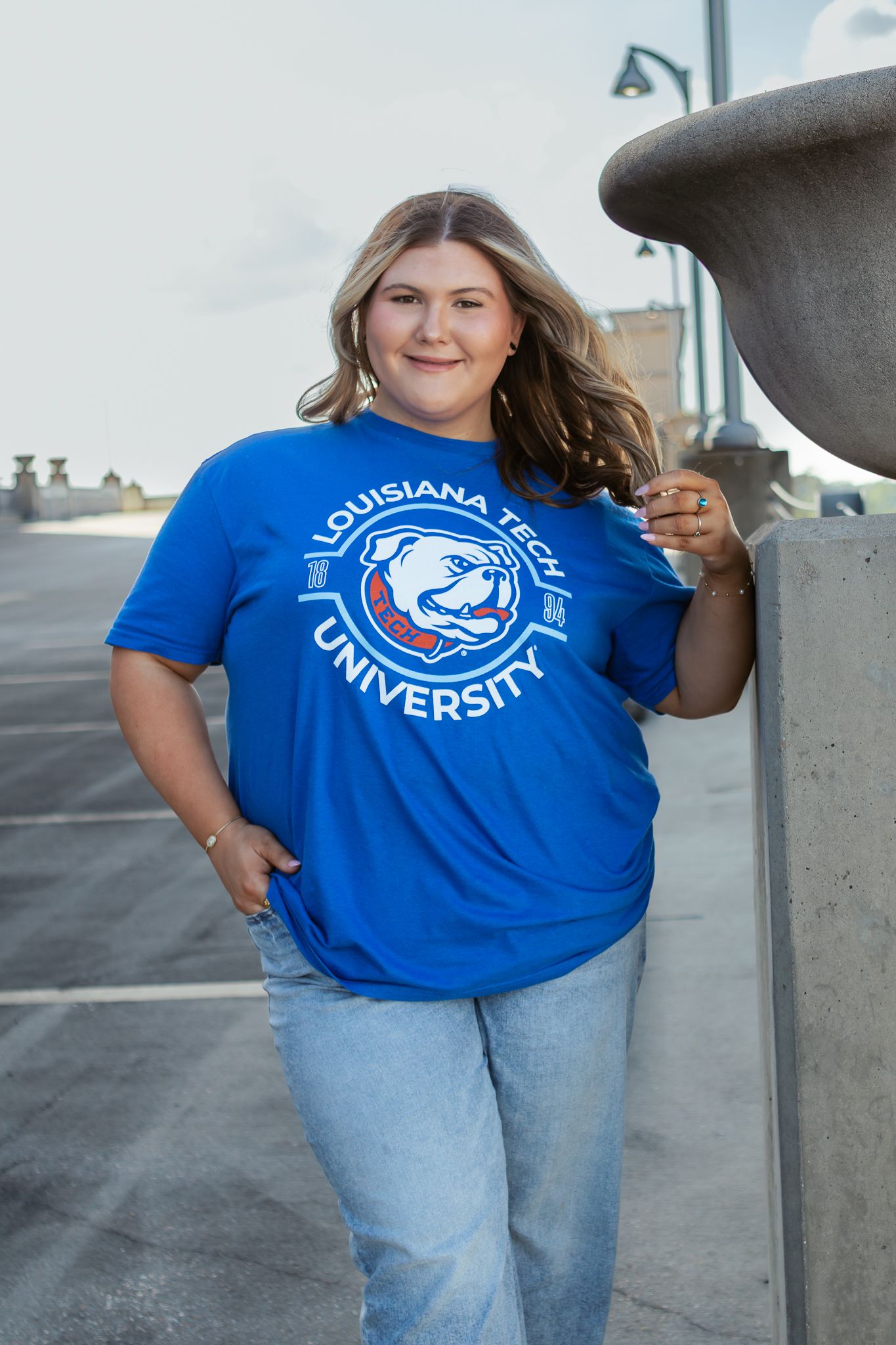 Smiling young woman wearing a royal blue Louisiana Tech University t-shirt and jeans leaning against a concrete pillar.