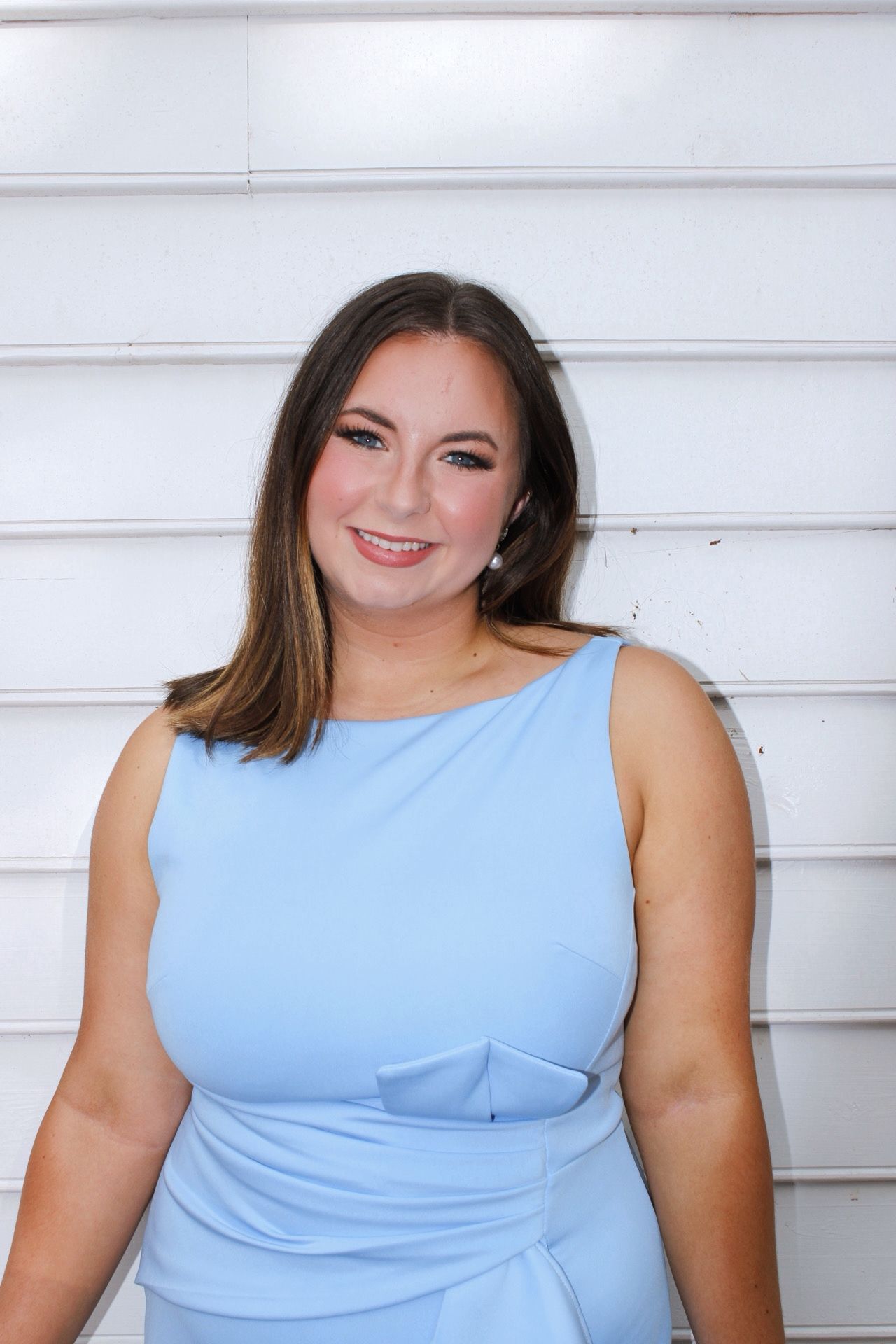 Smiling brunette woman in light blue sleeveless dress poses against white wall.