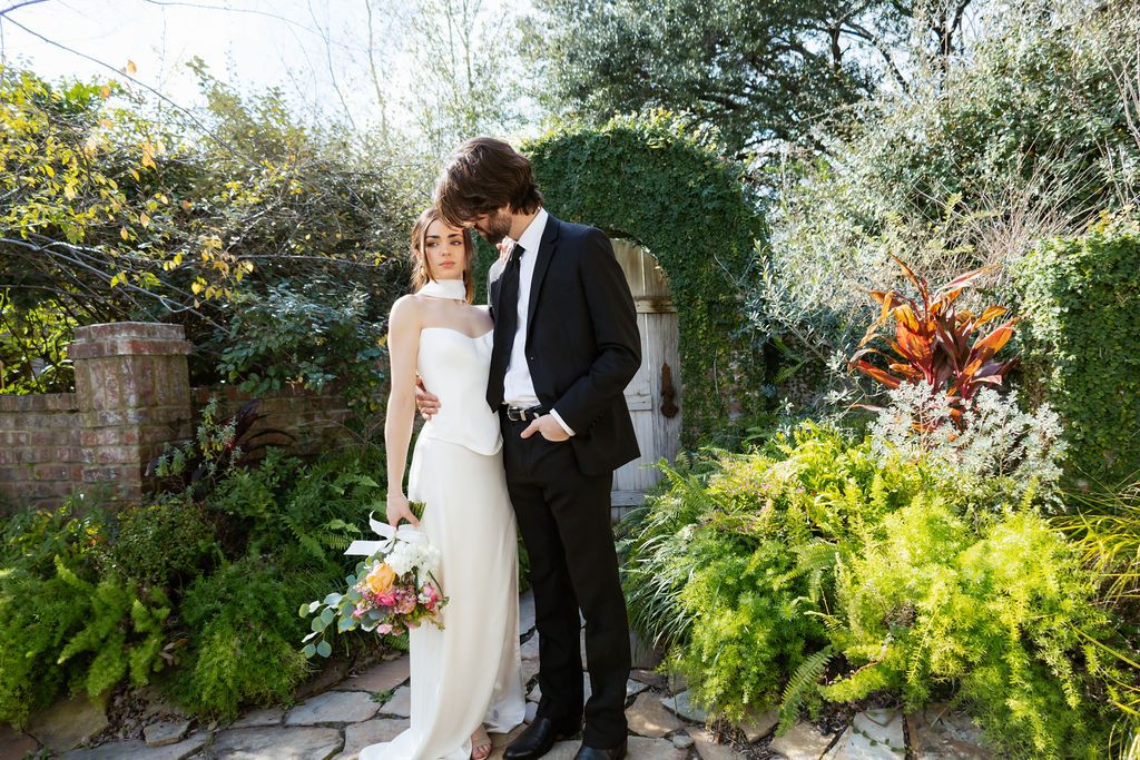 Bride and groom pose romantically in a lush garden setting, bride holding a colorful bouquet in a sleek white gown.
