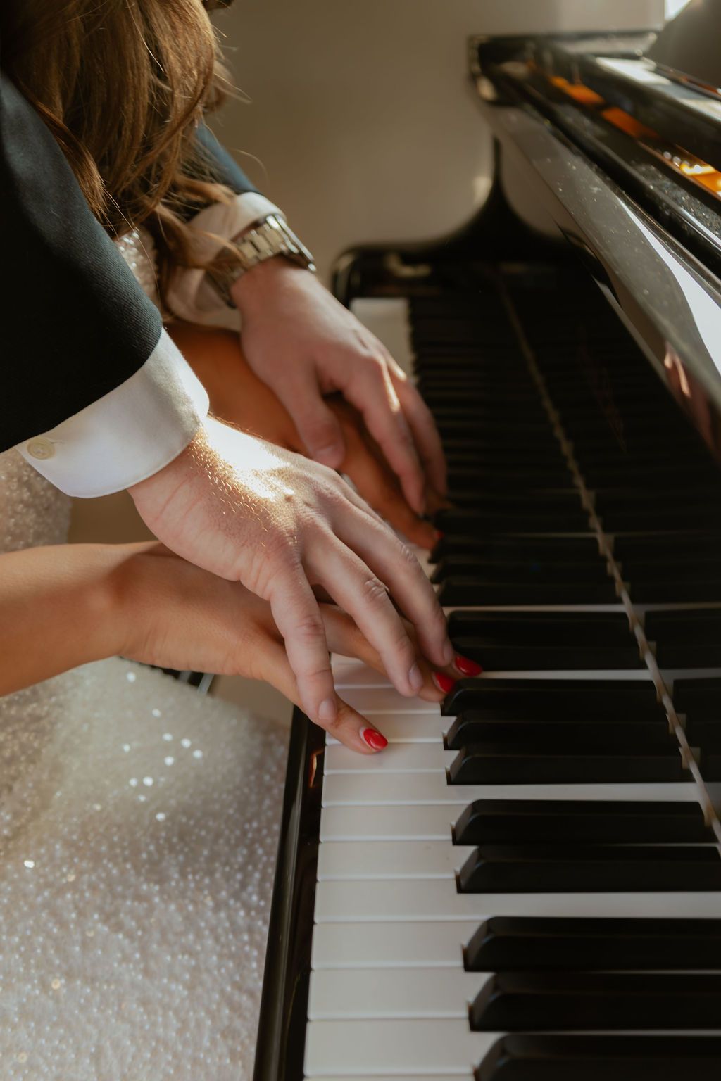 Two pairs of hands playing piano keys, one wearing red nail polish and sparkly dress.