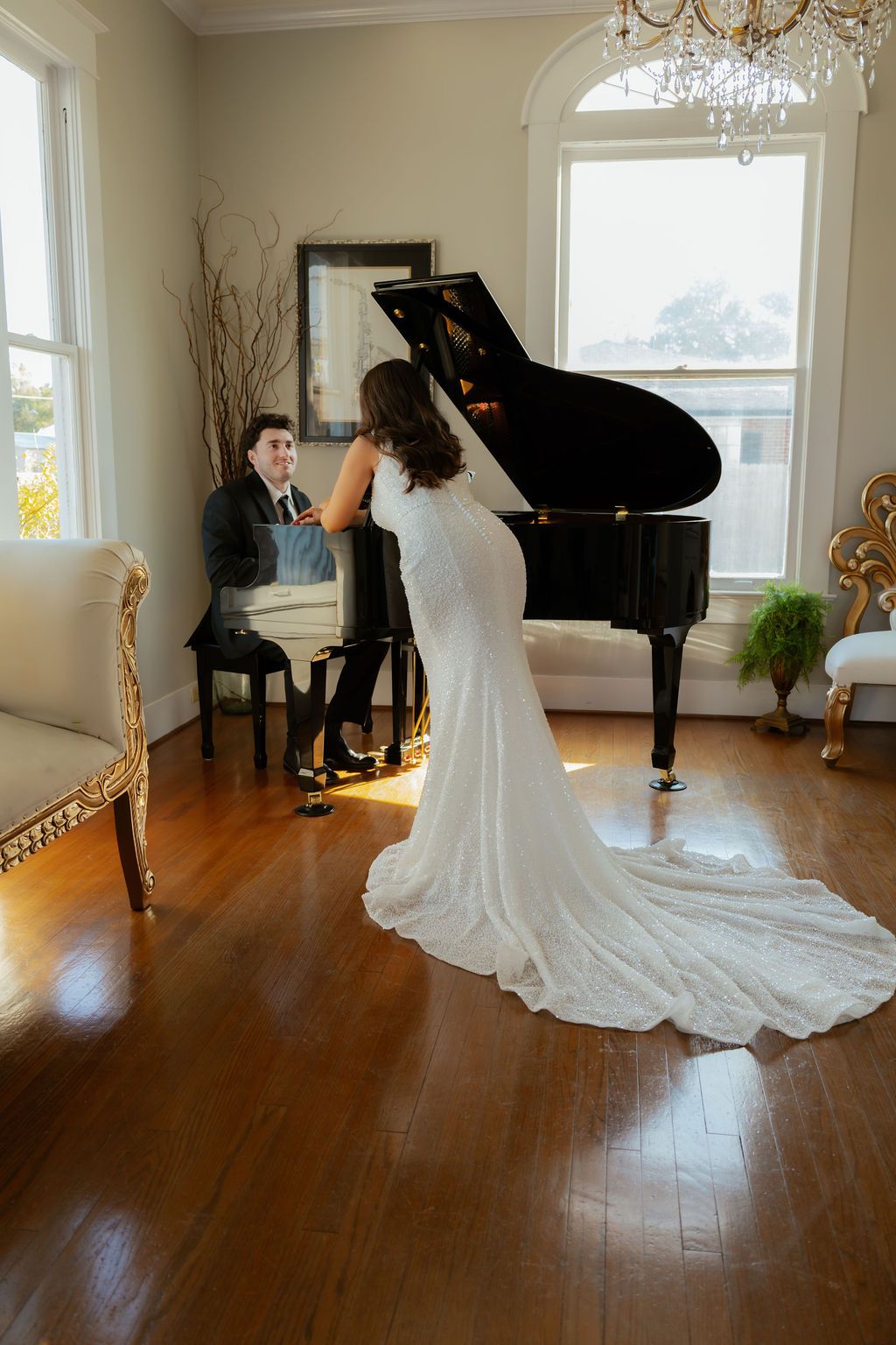 Bride in lace gown with train stands by black grand piano as groom plays.