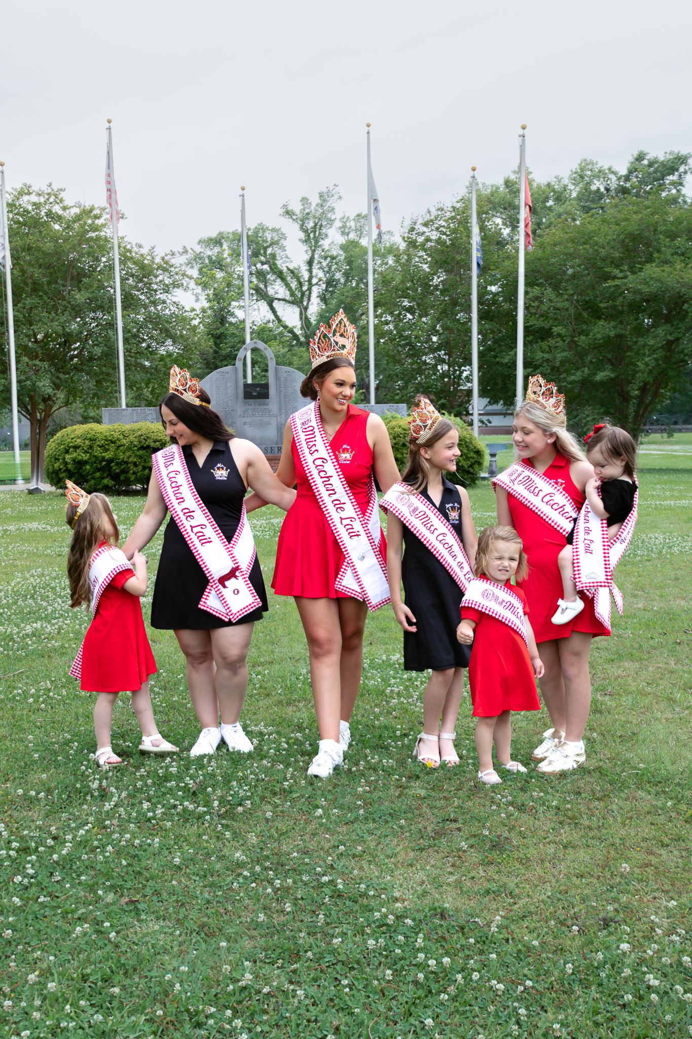 Pageant queens in red and black dresses with crowns and sashes walking in green park with flags.