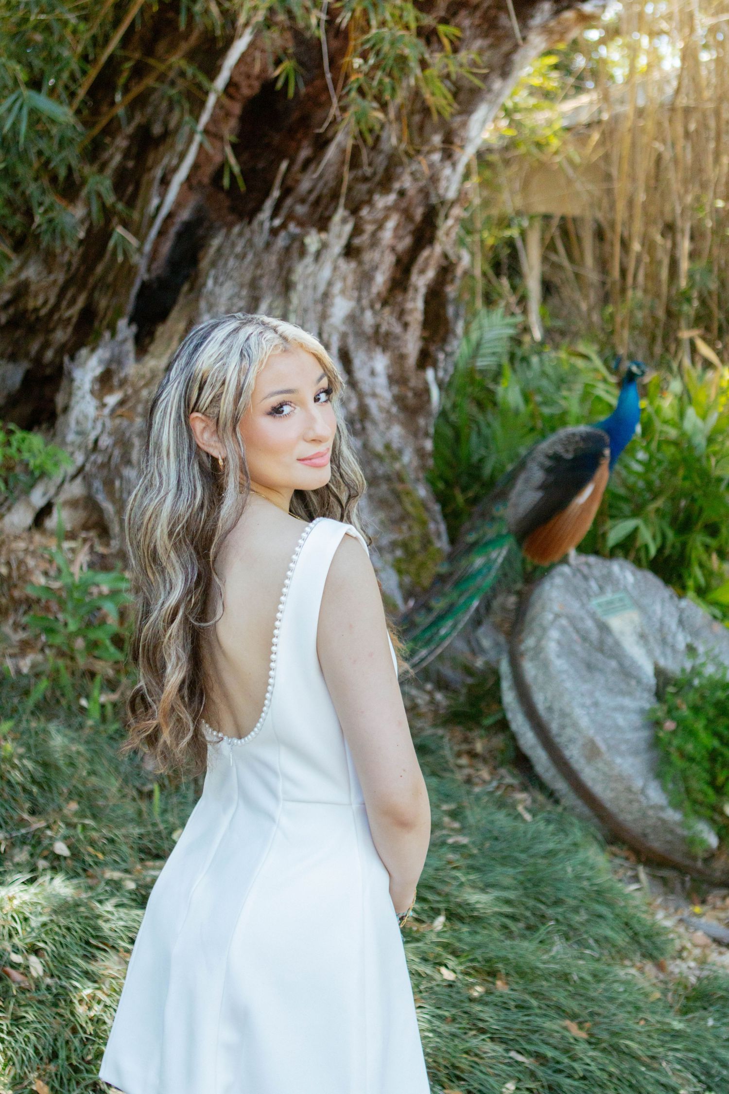 Elegant woman in white backless gown with peacock in lush garden background during golden hour.