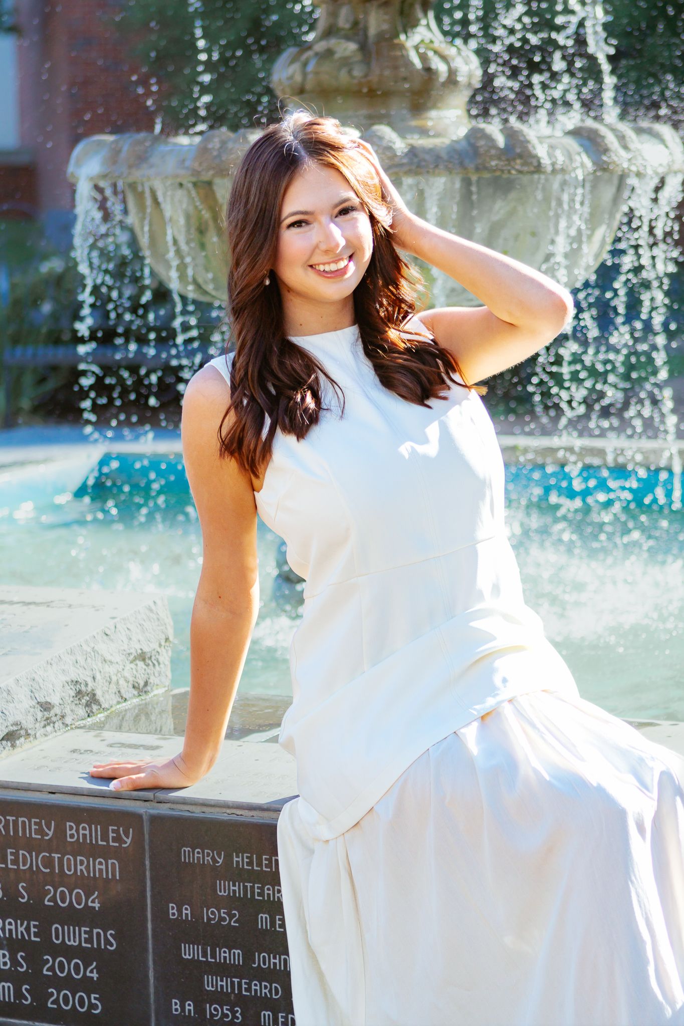 Smiling young woman in a white sleeveless dress sits by a stone fountain on a sunny day.