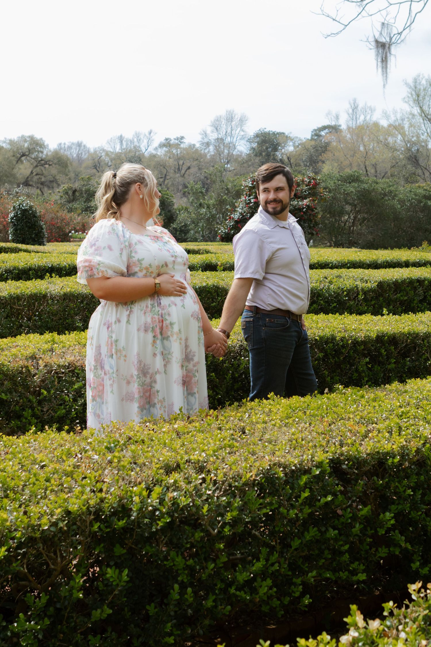 Pregnant woman in floral dress holding hands with bearded man in formal garden hedge maze.