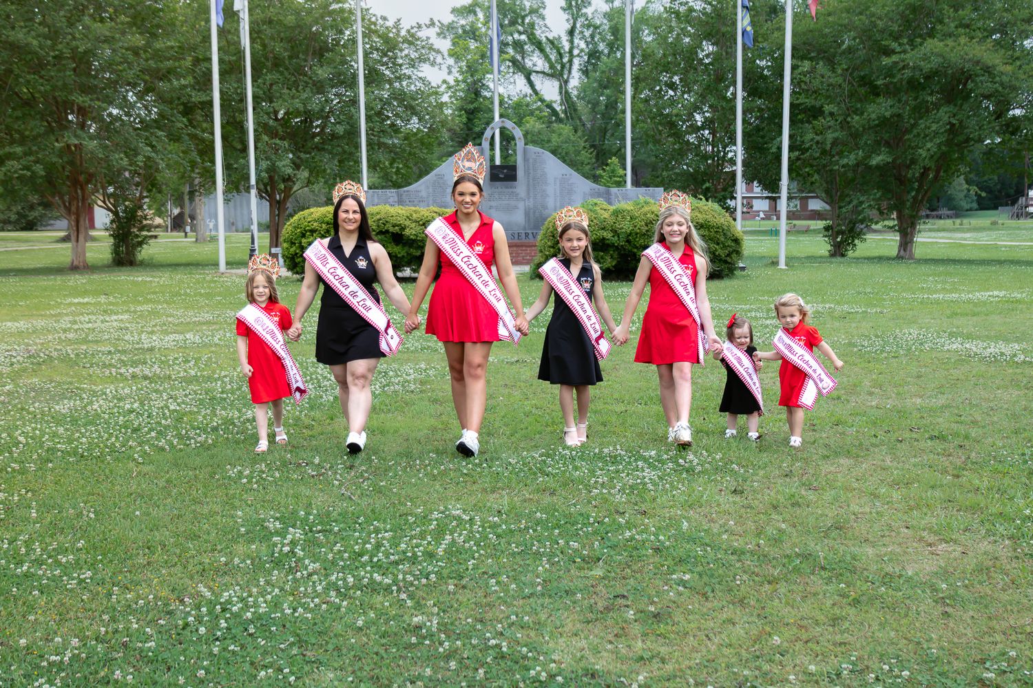Pageant queens in red dresses with crowns and sashes walking together across a green grassy field.