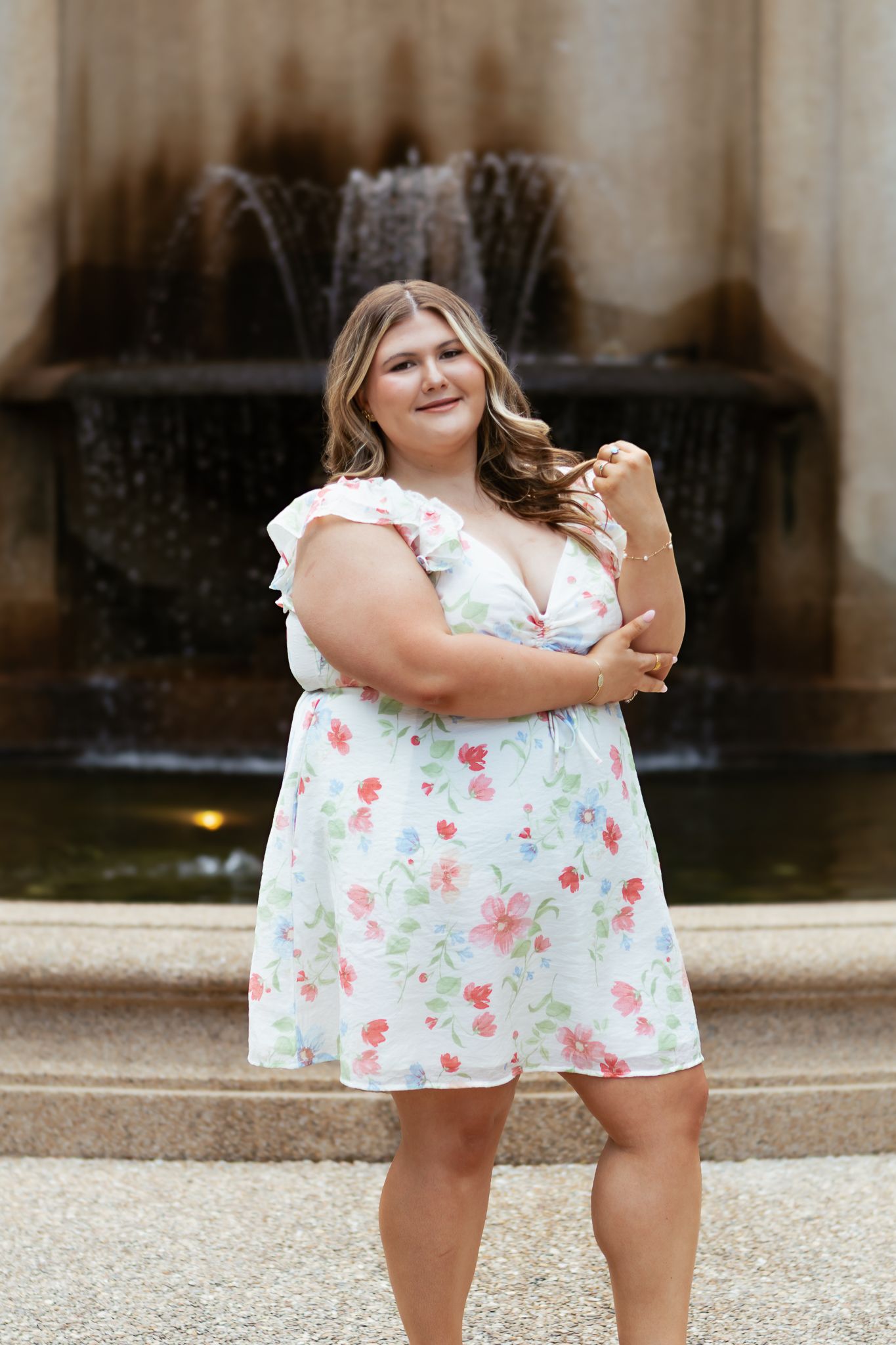 Smiling young woman in white floral dress standing in front of a decorative stone fountain outdoors.